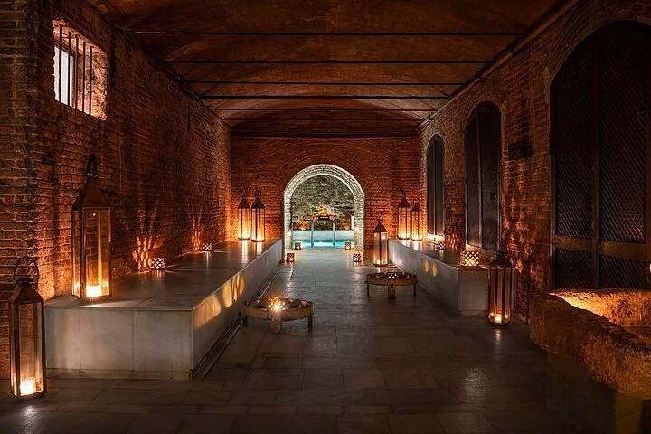 A brick-walled corridor with lanterns and candles, leading to an arch and outdoor pool area at night.