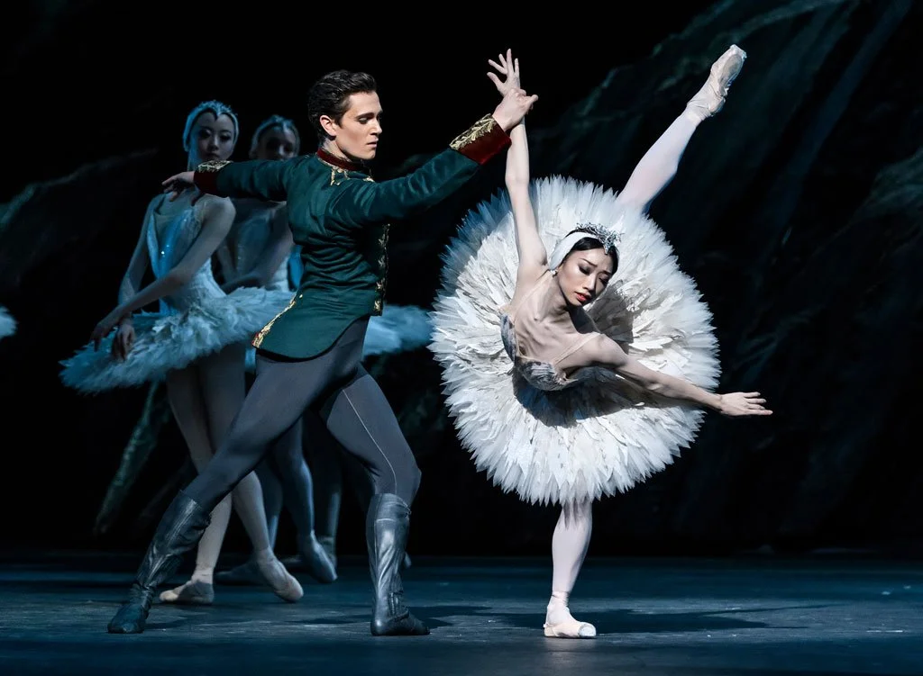 Ballet dancers performing on stage, with a male dancer in a green costume and a female dancer in a white tutu, in a classical ballet pose.