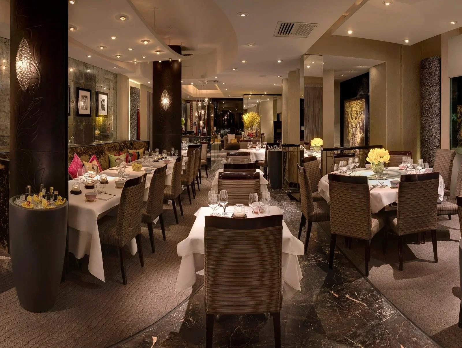 Elegant restaurant dining area with round and rectangular tables set with white tablecloths, glassware, and floral centerpieces, surrounded by striped chairs and decorative lighting, featuring marble flooring and artwork on the walls.