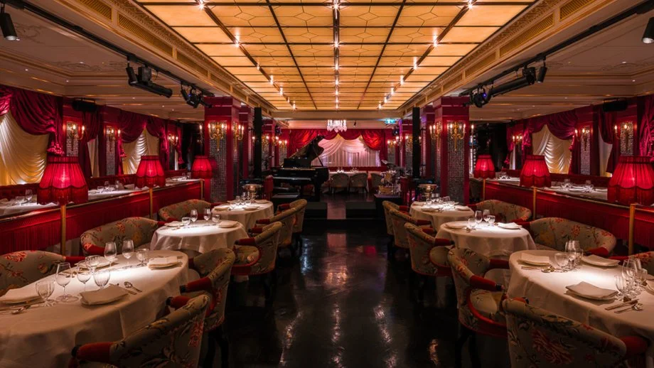 Elegant restaurant with round tables set with white tablecloths, wine glasses, and napkins. Red lamps and curtains decorate the space, with a grand piano at the far end and a stage with draped curtains and chandelier overhead.