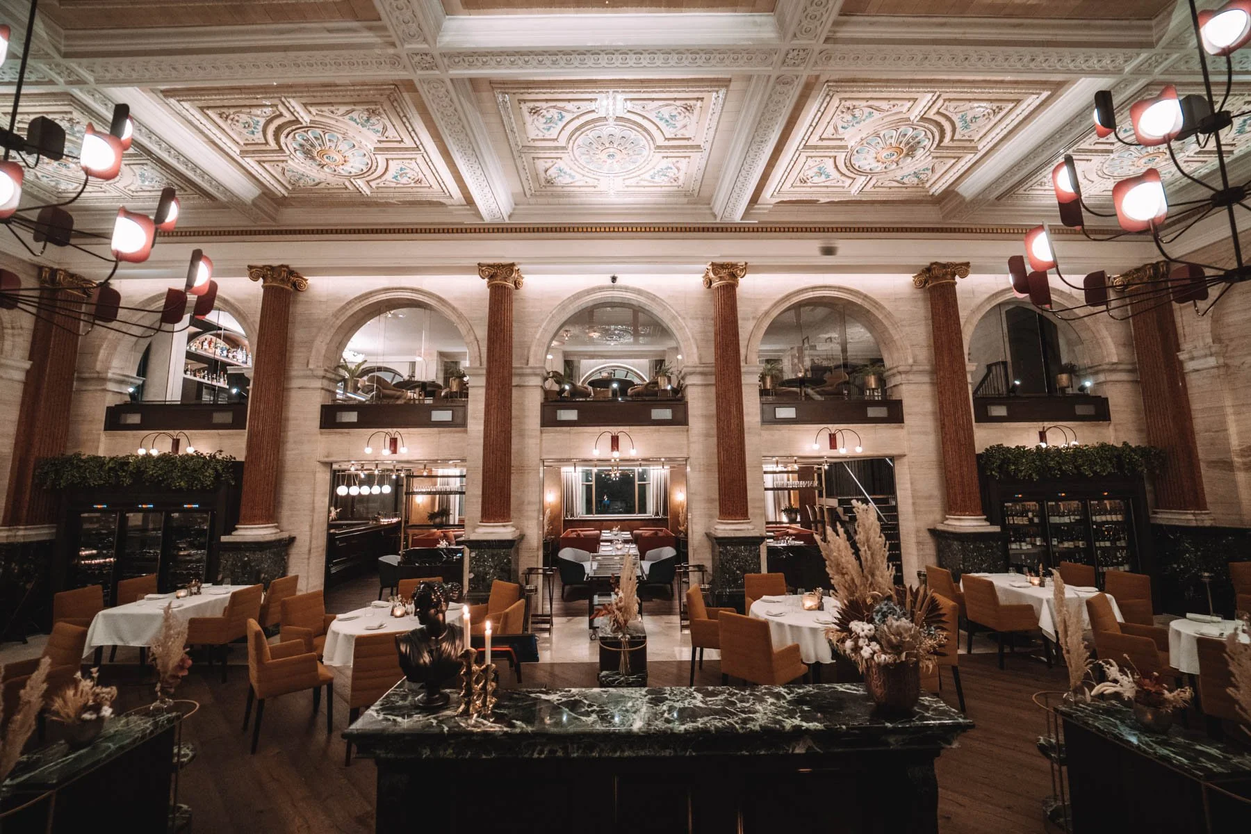 Elegant restaurant interior with high ornate ceiling, arched windows, and multiple tables with white tablecloths and candles, decorated with plants and vases.