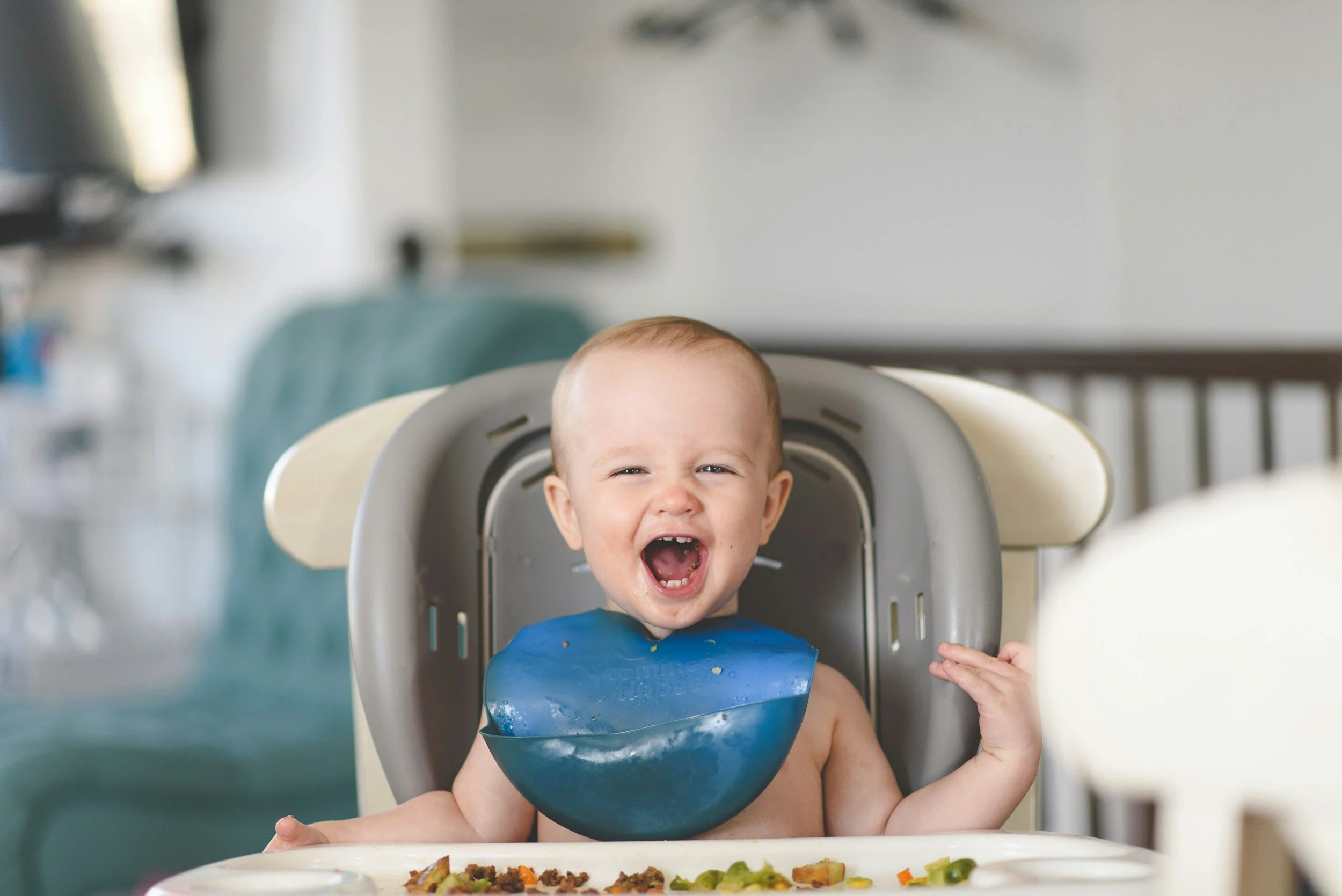A happy baby sitting in a high chair with a blue bib, laughing and enjoying a meal with food on the tray in front. The baby is starting solids.
