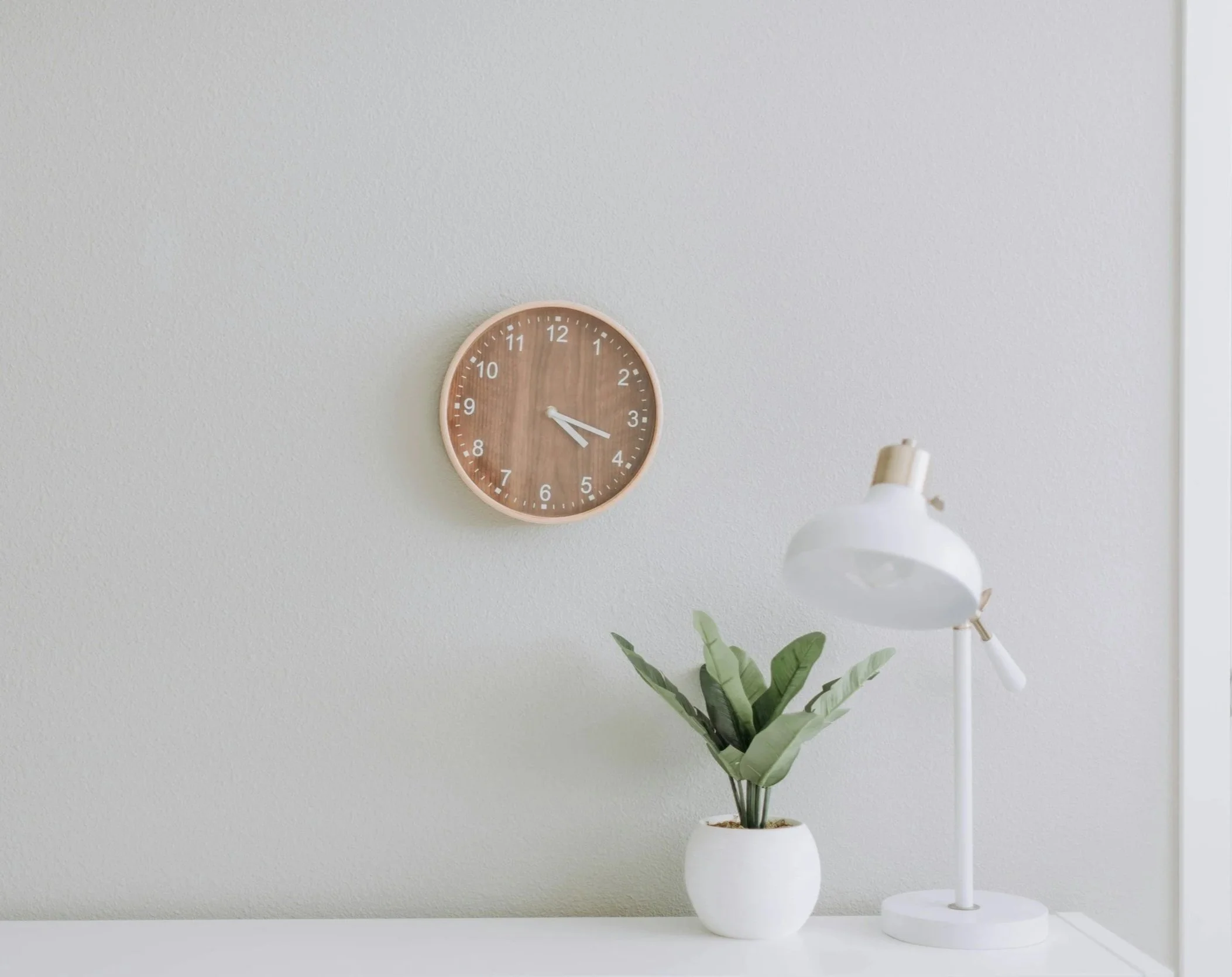 A wall clock with a wood face showing approximately 3:15, along with a white desk lamp and a potted plant with green leaves on a white surface against a plain, light-colored wall. Get in-office help from an IBCLC.