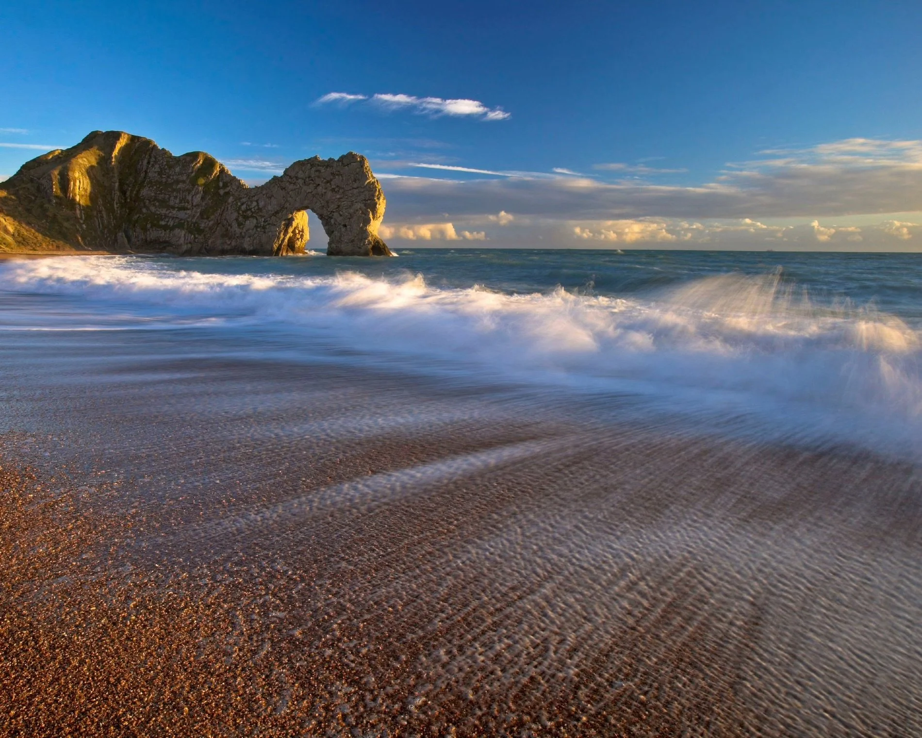 Waves breaking on a sandy beach at golden hour, with the Durdle Door limestone sea arch rising from the water beneath a blue sky.