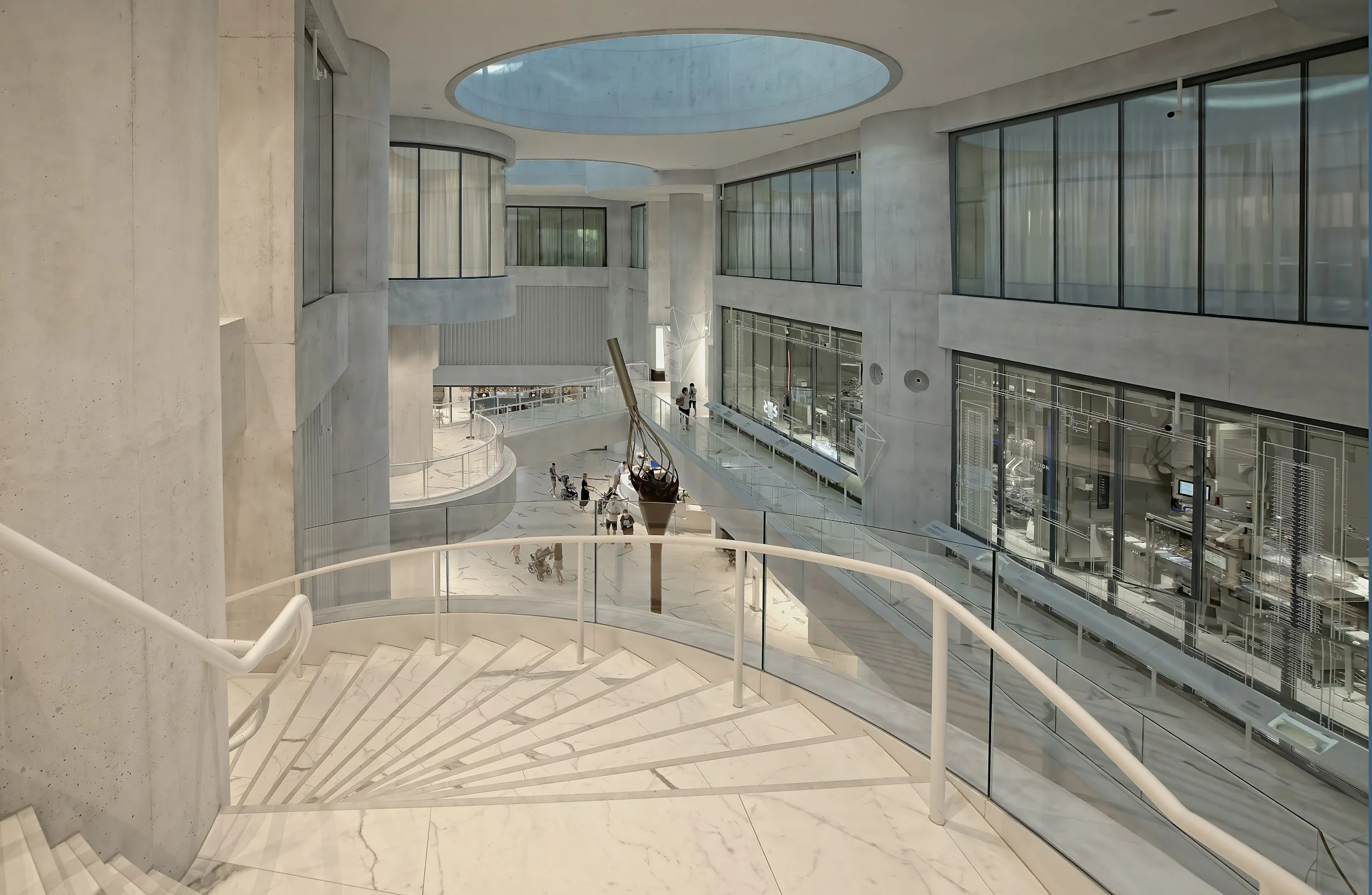Interior view of a modern building with white marble stairs, large glass windows, and a circular skylight overhead. People are walking in the lower level.