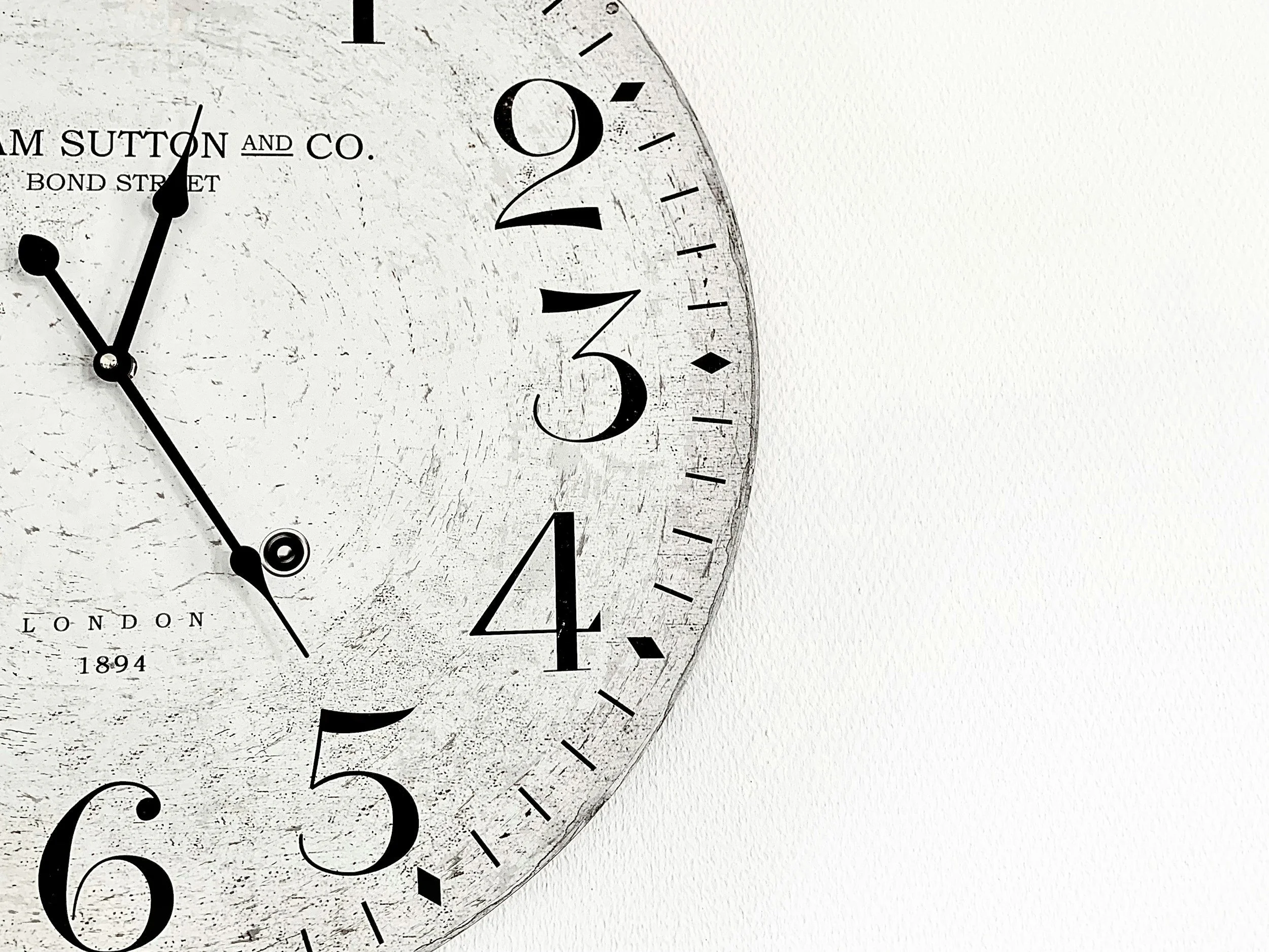 Close-up of a vintage-style clock with black hands and numbers on a white distressed face. The clock shows approximately 11:27.