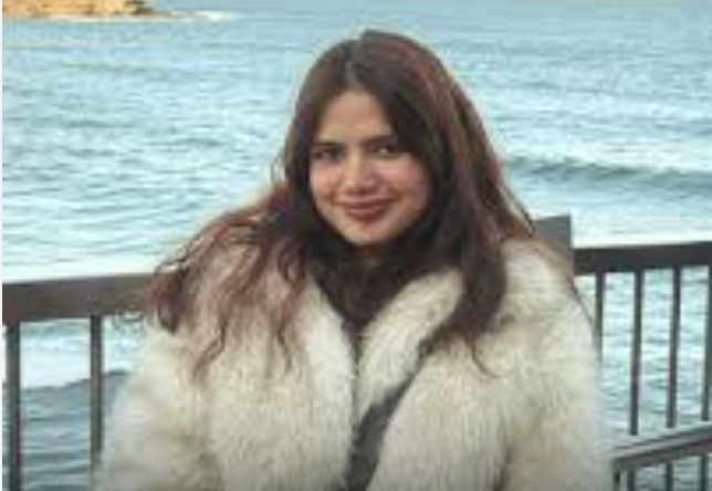 A young woman with long, wavy brown hair smiling, wearing a white fur coat, standing near a body of water with a wooden railing in the background.