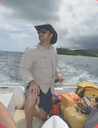 Person wearing a cowboy hat and sunglasses standing on a boat with a body of water and cloudy sky in the background, surrounded by yellow and orange bags and belongings.