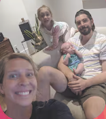 A family of four sitting together in a living room, smiling at the camera. The mother has blonde hair and is holding a baby, the father has a beard, and their teenage daughter stands behind them.