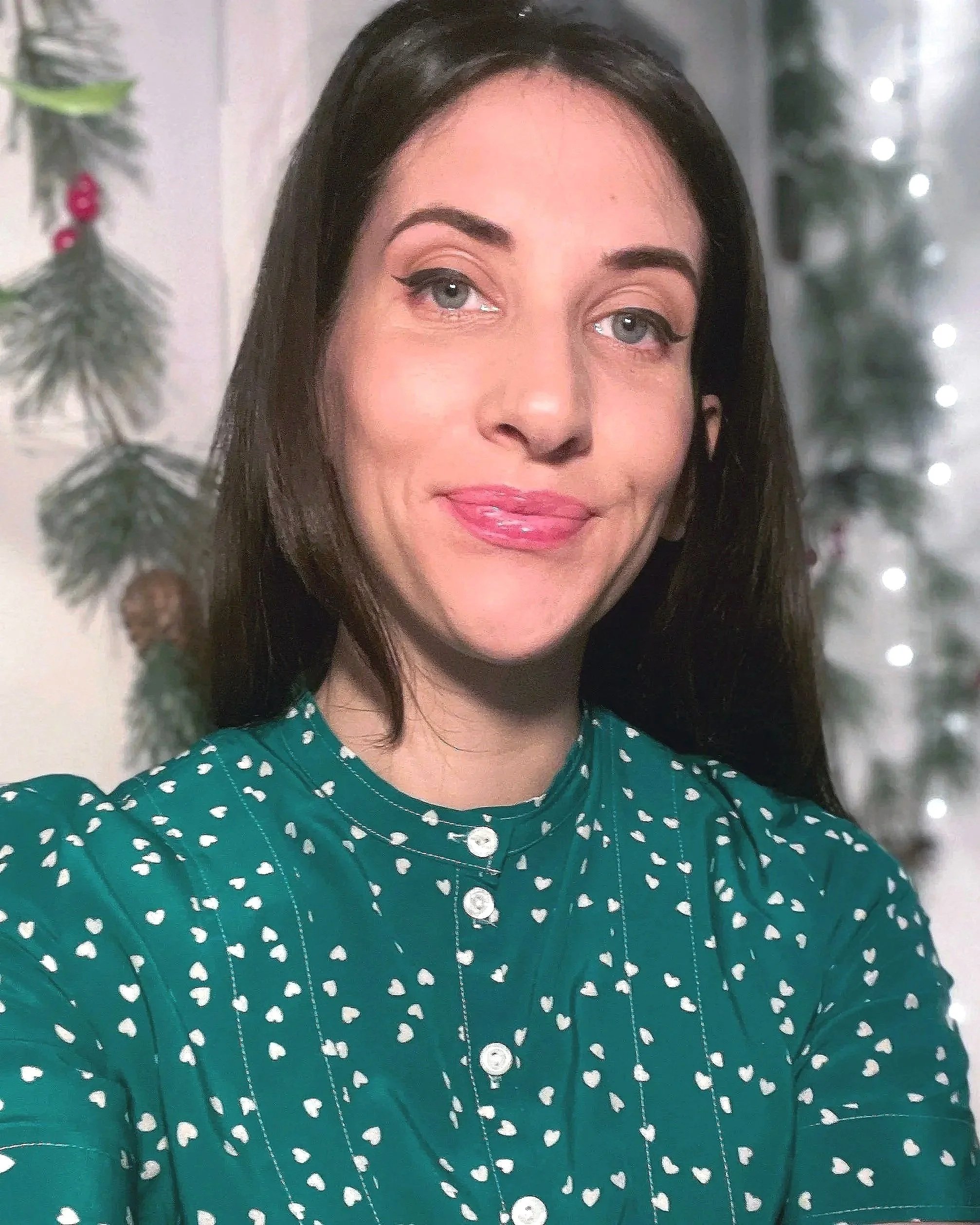 A woman with long dark hair and light skin, wearing a green top, smiling softly, with a blurred background of lights.