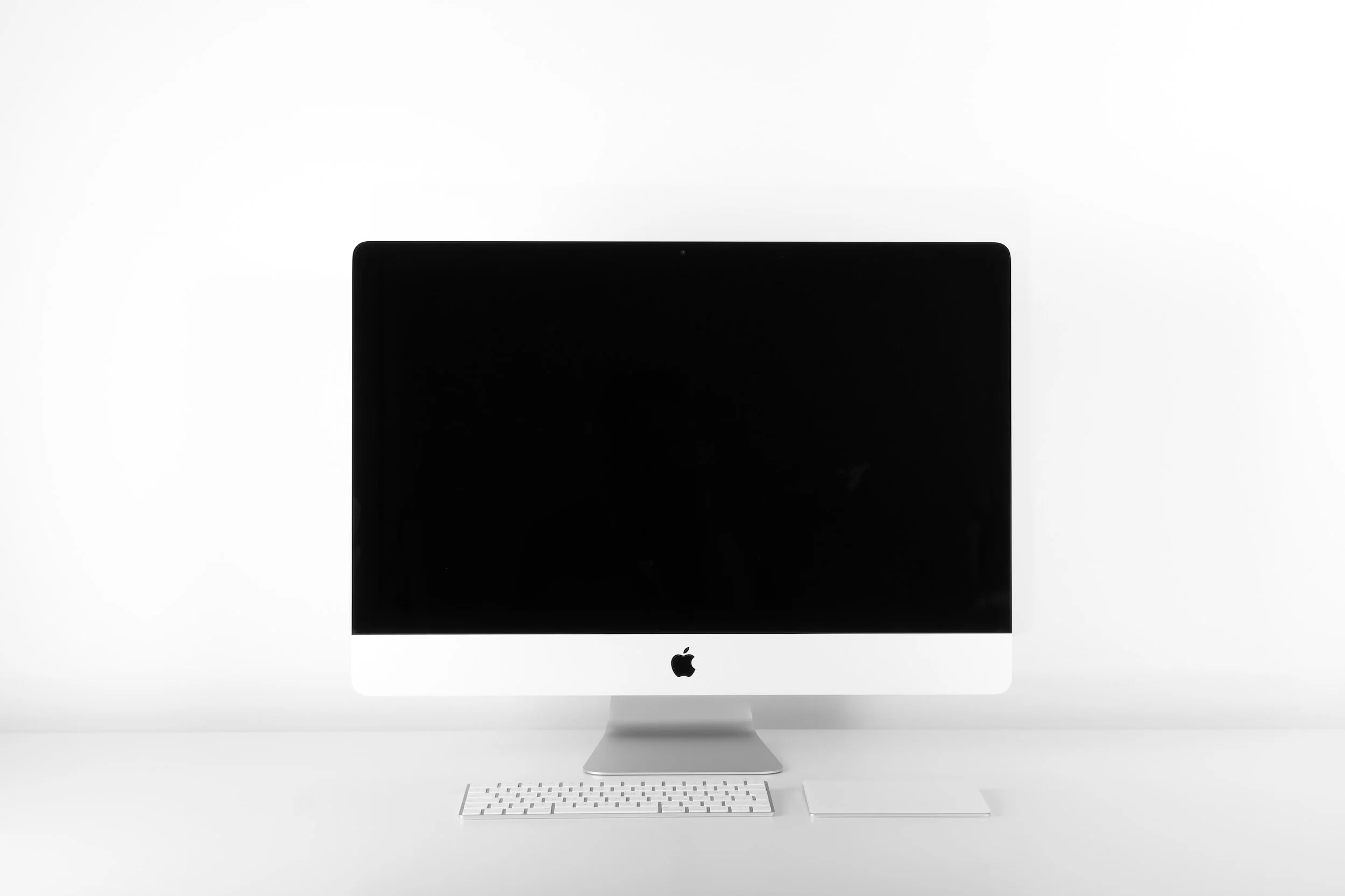 Apple iMac desktop computer with a black screen, white keyboard, and a white trackpad on a white background.
