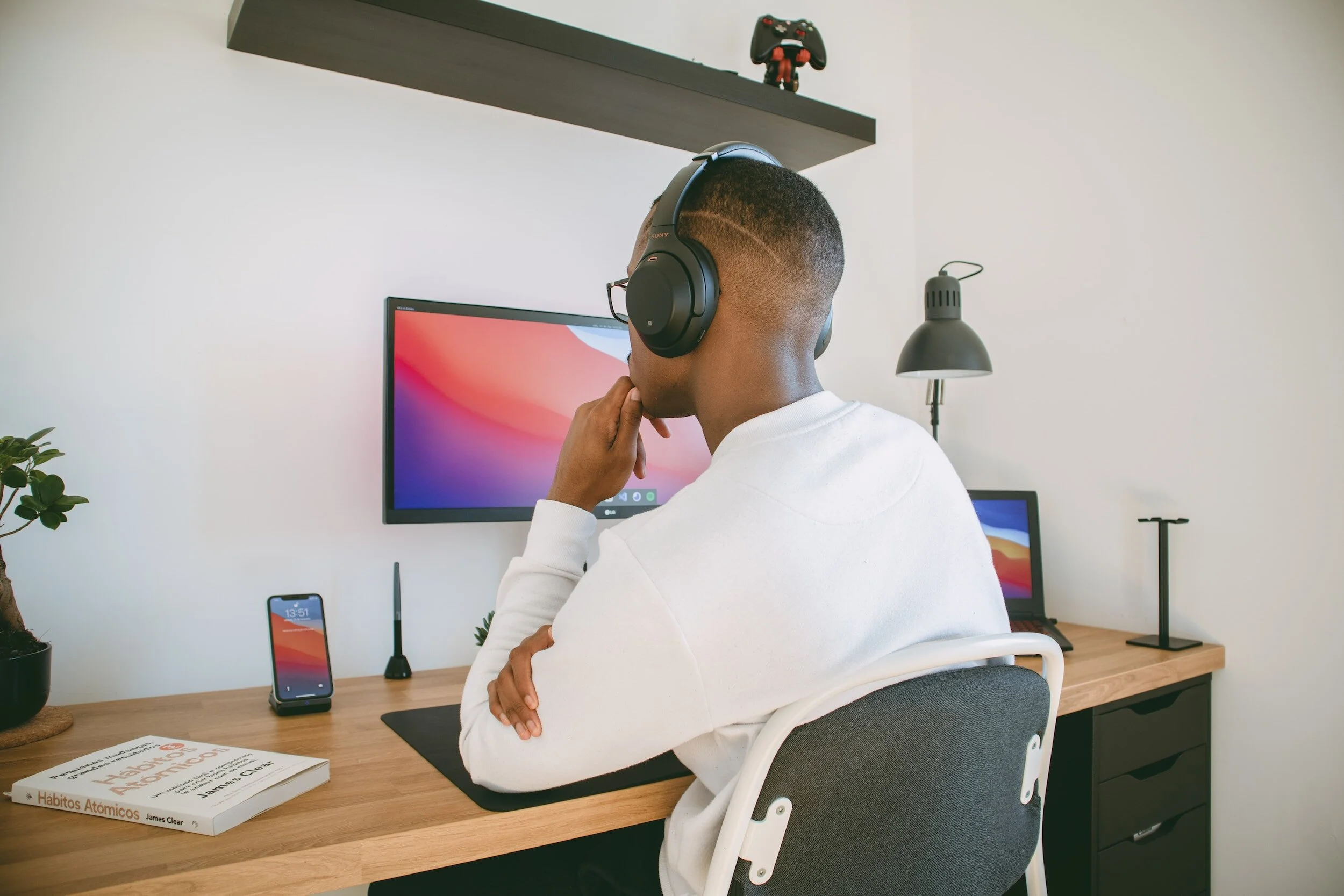 A person wearing headphones sitting at a desk with a computer monitor, a laptop, a smartphone, a book, and a desk lamp in a modern room.