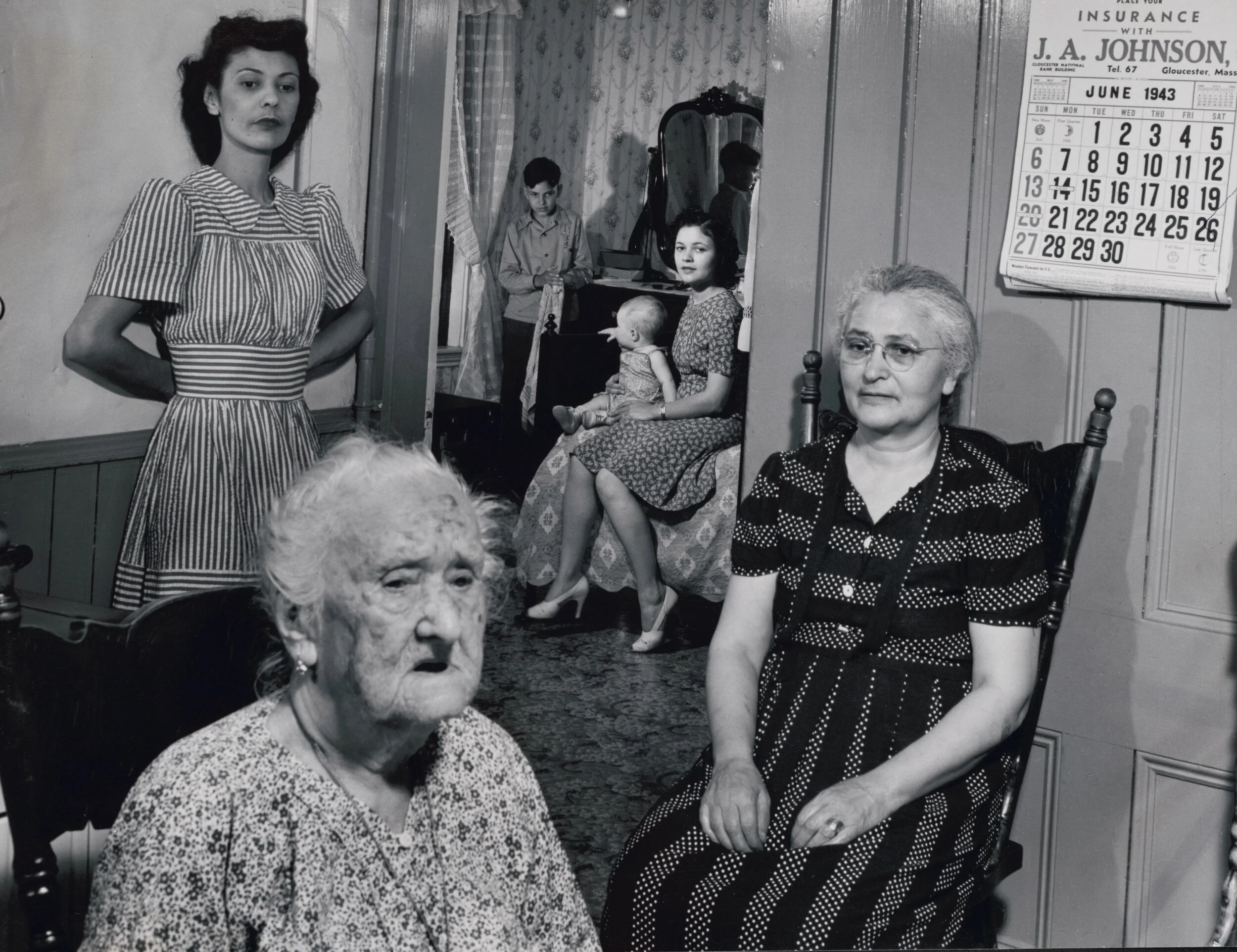 A black and white photo of five women and two children in a room, with a wall calendar showing June 1943. The women are seated or standing casually, with one woman in the foreground with a surprised or shocked expression. The background includes a woman sitting with a toddler on her lap, a standing boy near a window, and a woman with dark hair and a striped dress standing with her hands on her hips.