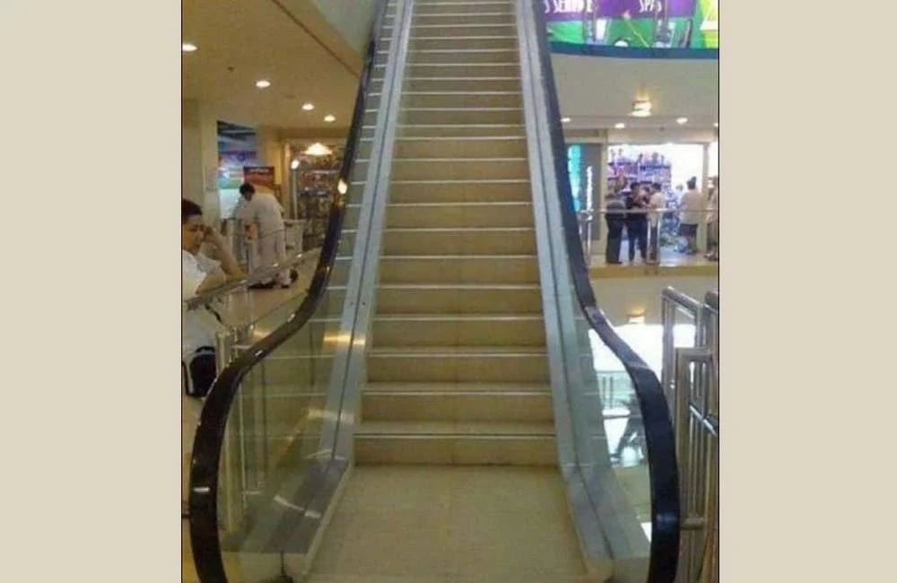Escalator in a shopping mall with a staircase placed upside down on top, creating an optical illusion.