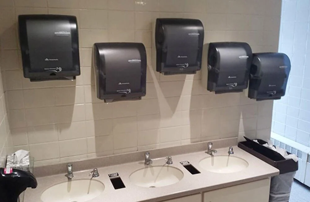 Five black paper towel dispensers are mounted on a beige tiled wall above a beige sink countertop with three sinks. There is a black trash bin on the right side and some items on the left side.