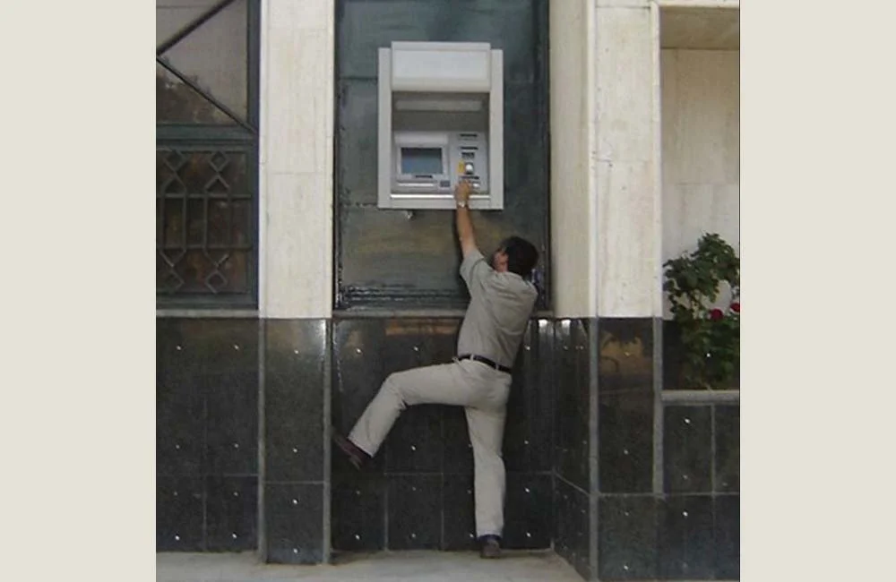 A man in a gray shirt and light-colored pants appears to be hanging onto an ATM or bank kiosk, with one foot on the wall and one arm reaching toward the machine, creating the illusion that he's climbing or hanging on the ATM.