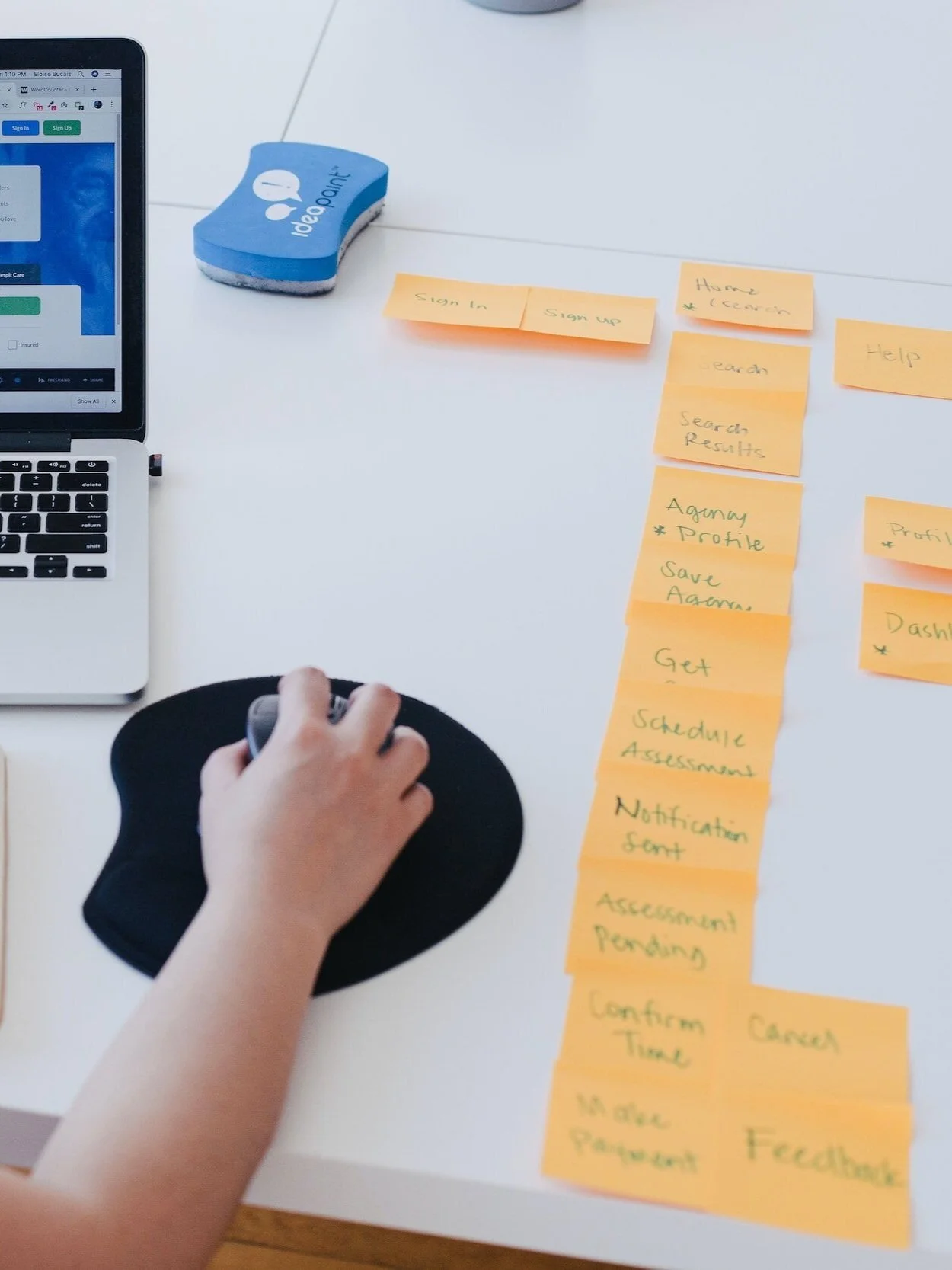 Desk with a laptop, sticky notes arranged in a workflow, a blue eraser, and a person's hand using a mouse on a black mouse pad.