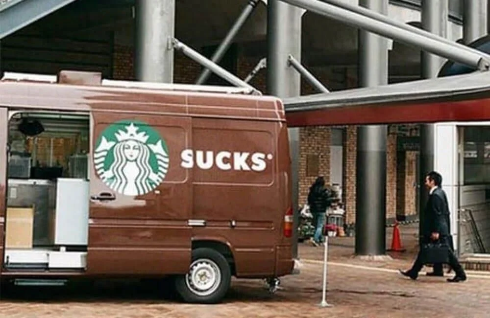 A brown Starbucks delivery van with the famous logo, but with the word 'SUCKS' instead of 'Starbucks'.