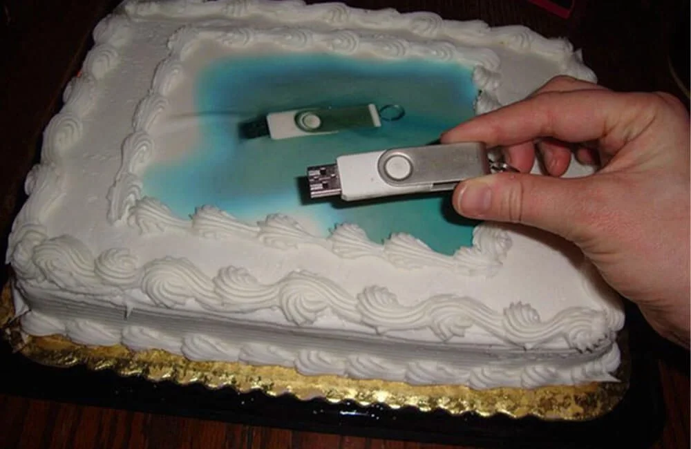 A square birthday cake with white frosting and piped borders, designed to look like a swimming pool with blue fondant in the center. A hand is holding a USB flash drive, which is being used to create a digital laser-engraved image of the same USB drive on the cake's surface.
