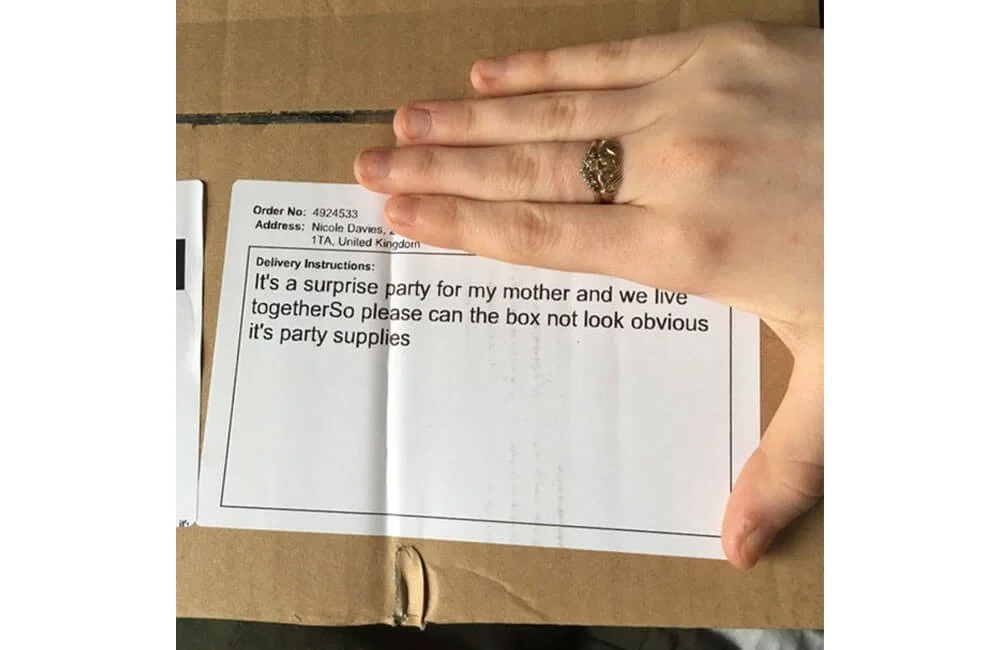 A hand with a ring on the ring finger resting on a cardboard box, with a note inside the box that says it's a surprise party for someone's mother, requesting the box not look obvious and asking for party supplies.