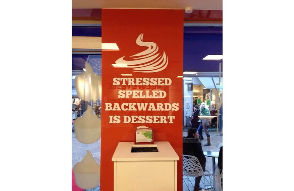 A sign inside a café with a logo of a steaming cup of coffee or tea. The sign reads, "STRESSED SPELLED BACKWARDS IS DESSERT." There is a small counter or display below the sign, with a napkin holder. The background shows a shopping mall or similar indoor space with people walking and sitting.