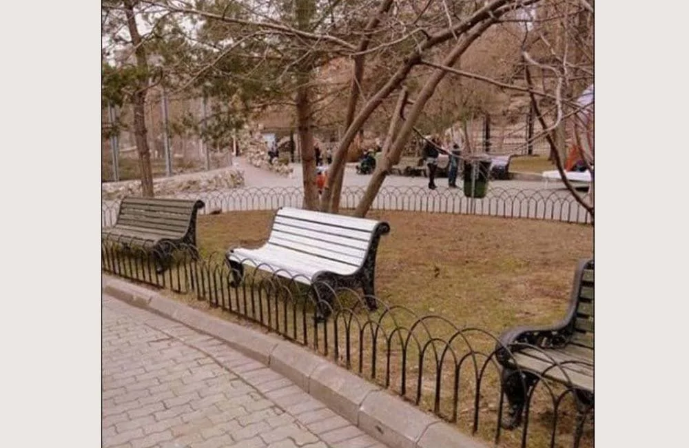 Empty park benches behind a small decorative black metal fence, with leafless trees and a pathways in the background.