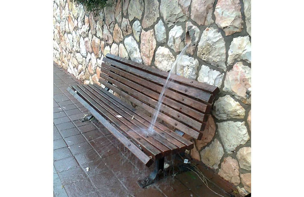 A wooden bench attached to a stone wall with a small water fountain spouting water onto it.