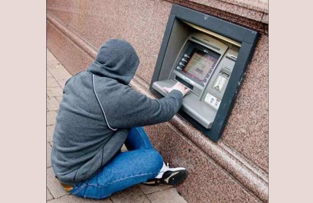 A person with a hooded sweatshirt and jeans using an ATM machine embedded in a brick wall outside.