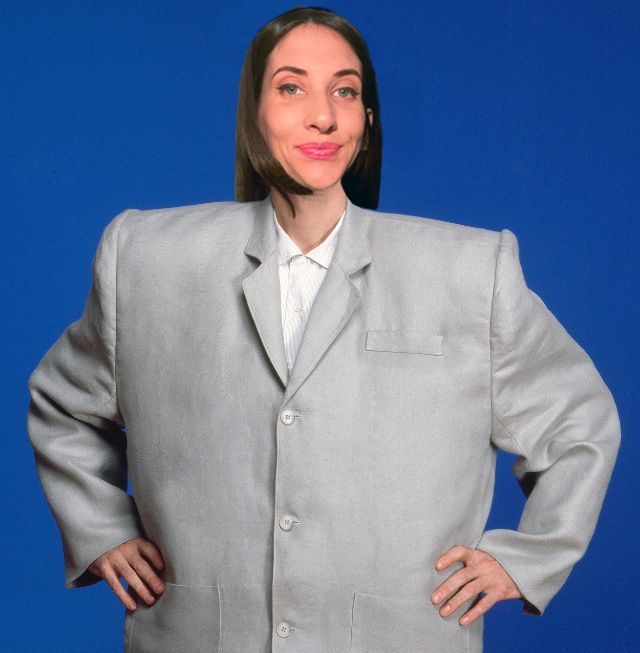 A woman with shoulder-length dark hair wearing a grey suit with wide shoulders and a white blouse, standing against a solid blue background, with a confident expression and hands on her hips.
