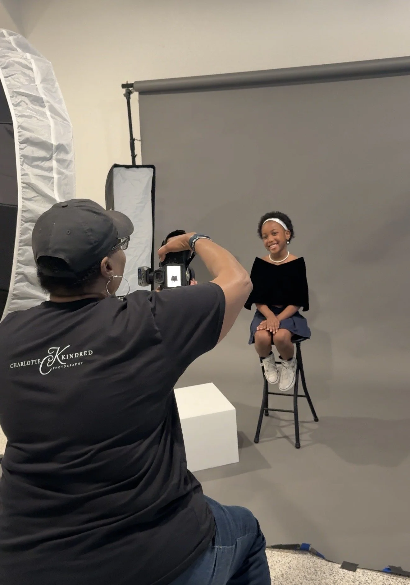 A woman taking a photograph of a young girl sitting on a stool in front of a gray backdrop. The girl is smiling, wearing a black top, blue skirt, white sneakers, a white headband, and pearl earrings.