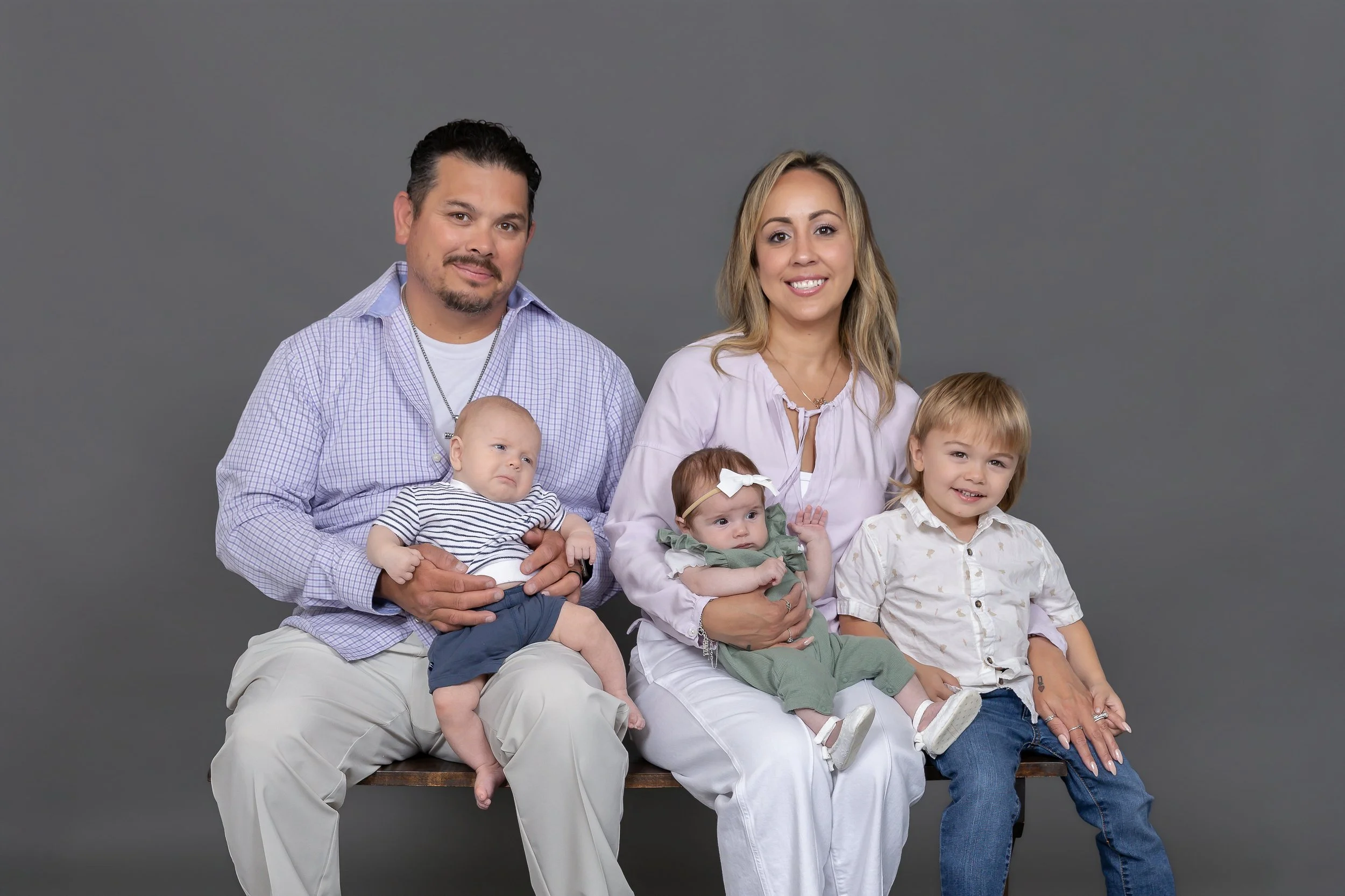 A family portrait with two parents and four children posed against a gray background. The father, with dark hair and a goatee, is sitting on the left holding a baby dressed in a striped shirt. The mother, with blonde hair, is sitting on the right hol