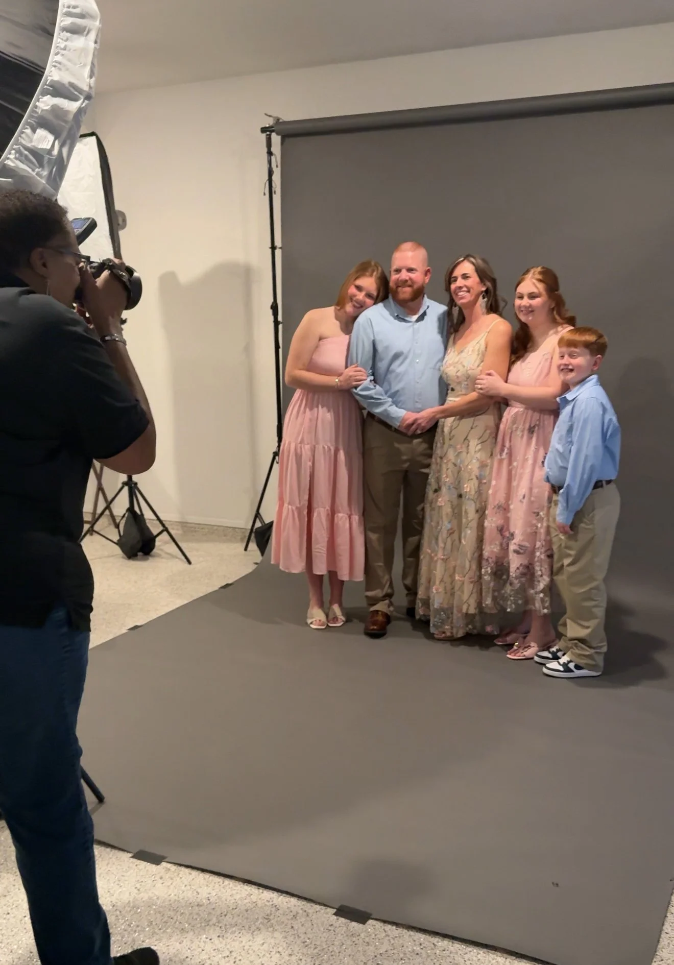 Group of five people, three women and two men, standing together in front of a grey backdrop in a photo studio. A photographer is taking their picture with a camera.