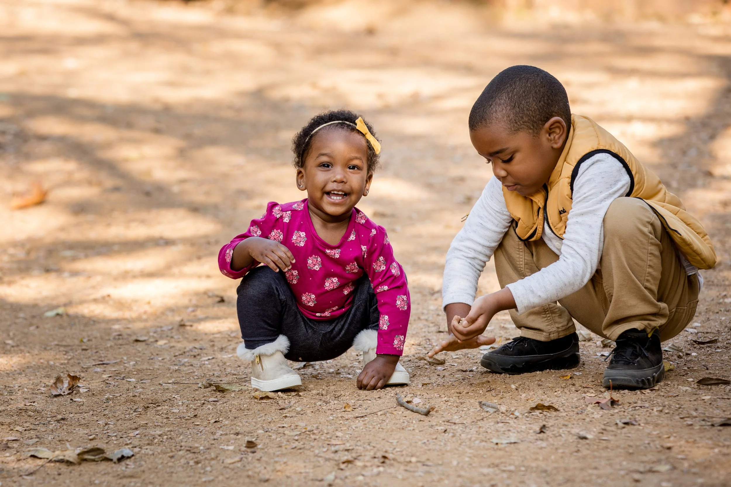 African-American little boy and toddler girl sitting and playing on a gravel trail in a park.