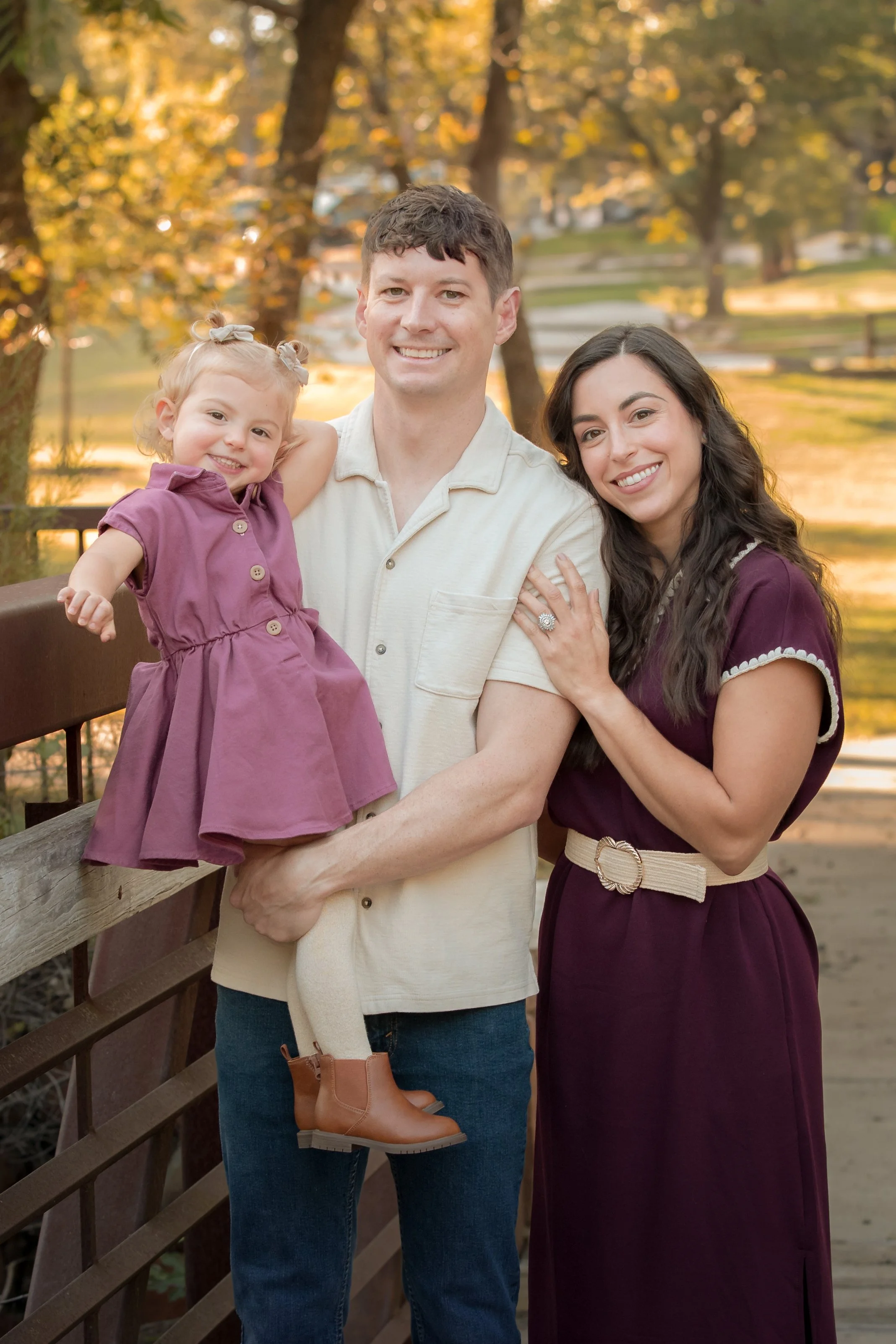 Family of three wearing coordinating cream, burgundy, and mauve outfits while posing together outdoors in the fall.