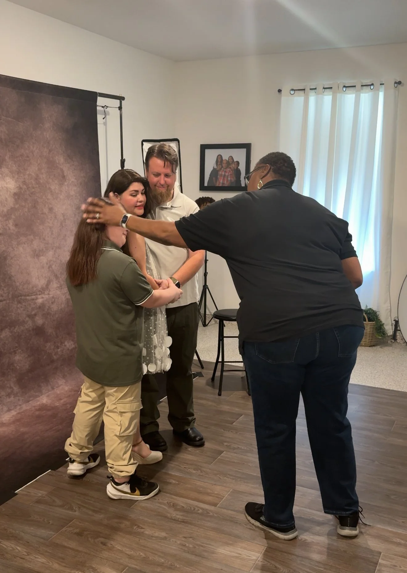 Four people gathered in a room, two of them are standing closely with one person touching the other's face gently, and the woman on the left appears emotional. Background includes a brown backdrop, a framed photo on the wall, a black chair, and white curtains covering a window.