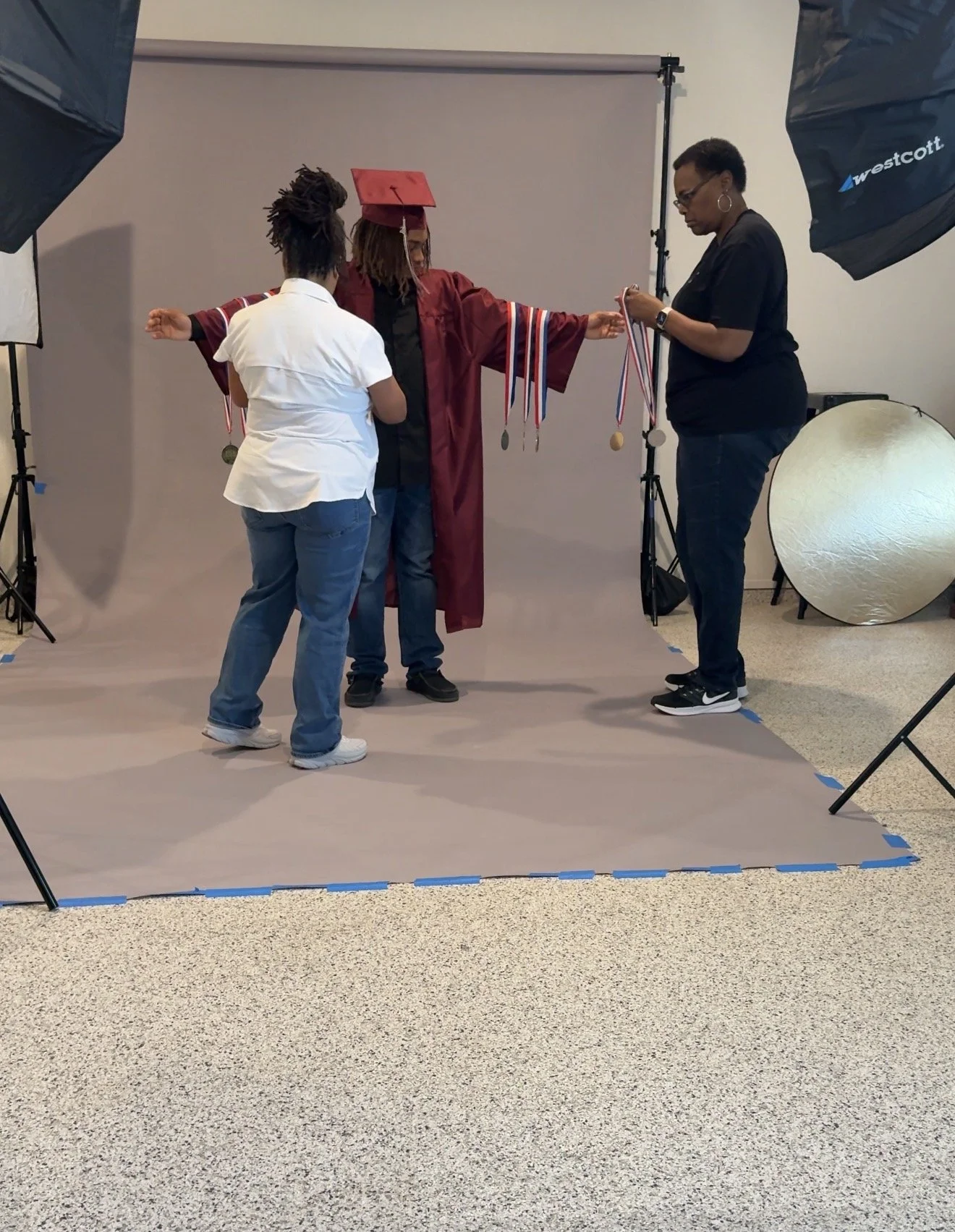 A graduation ceremony where a graduate in maroon cap and gown is receiving medals from a woman, with another woman standing nearby, in a photography studio with lighting equipment and a backdrop.