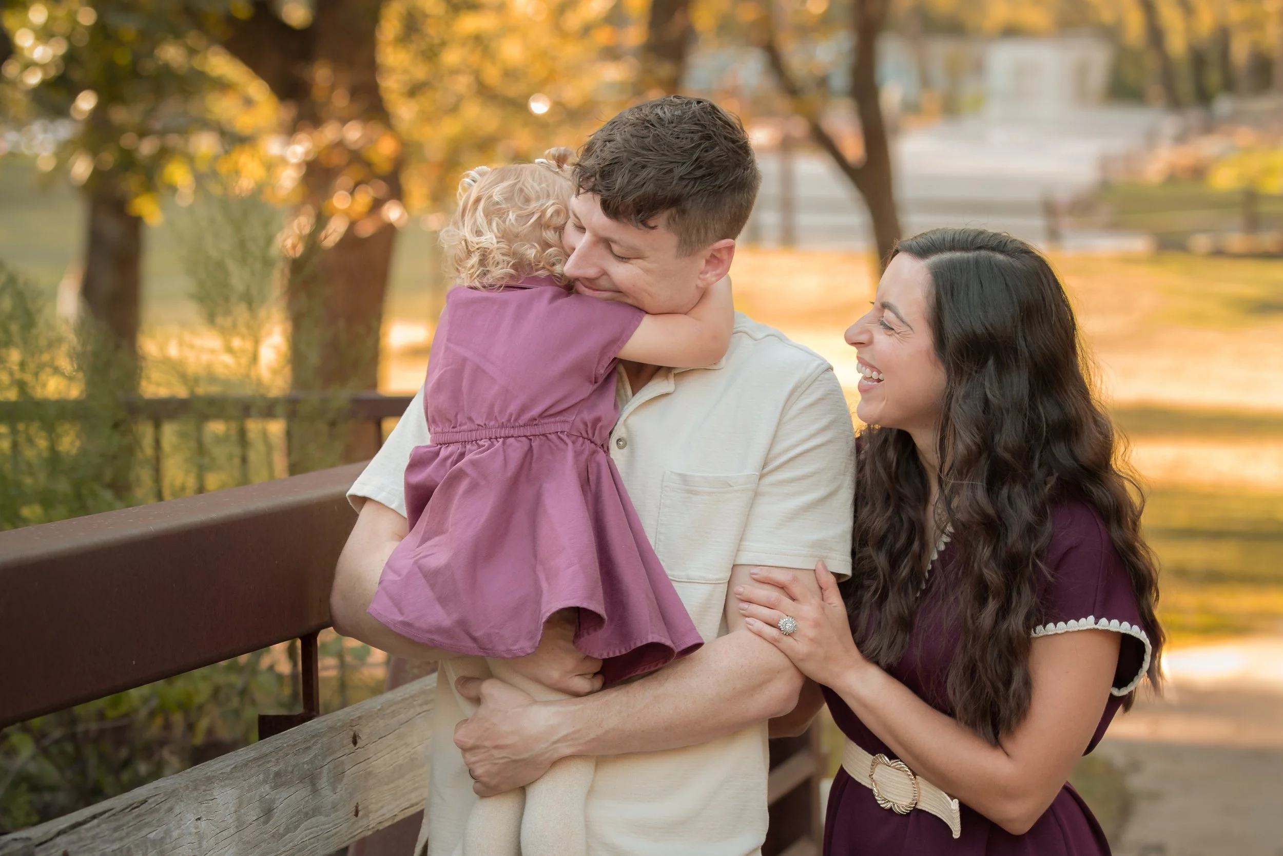 Young girl wraps her arms around her father's neck while her mother smiles beside them during an outdoor family session.