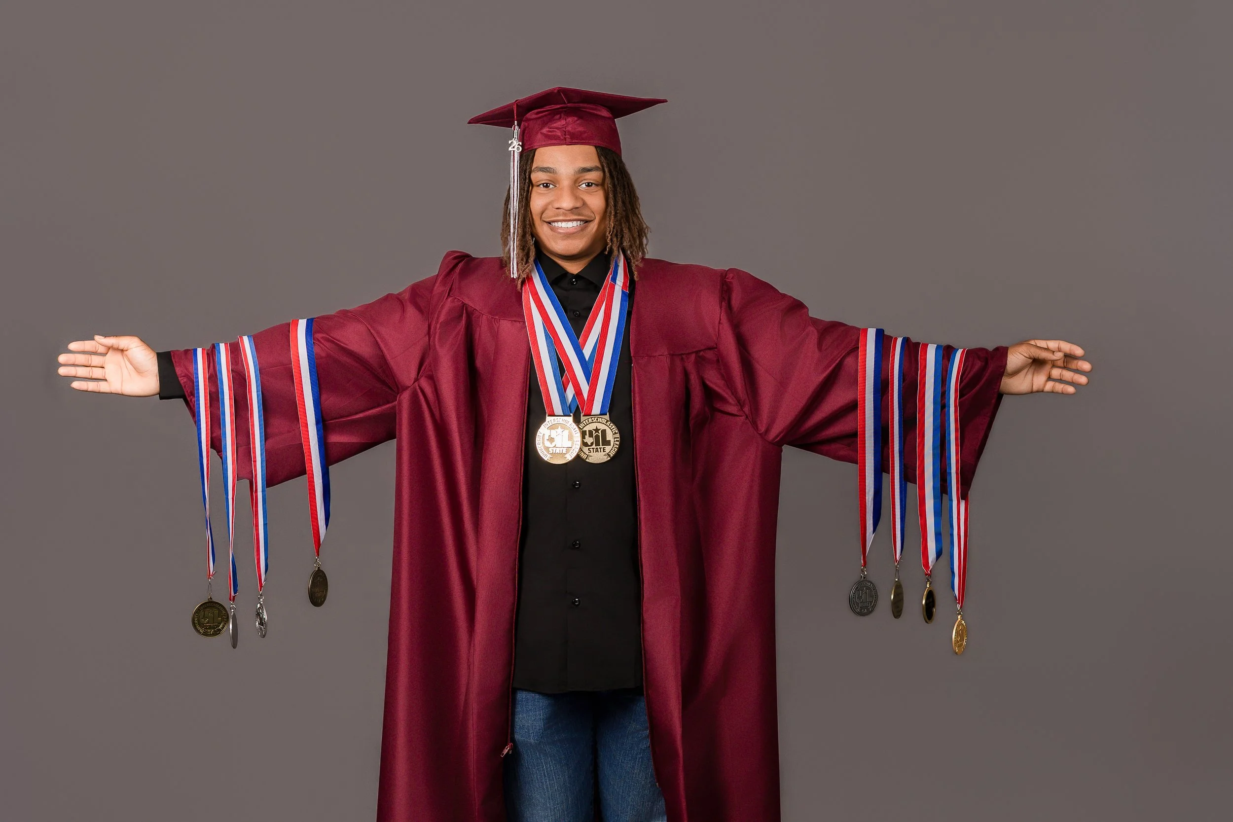 A person in a maroon graduation gown and cap, smiling and extending both arms outward, with medals hanging from ribbons around their neck against a gray background.