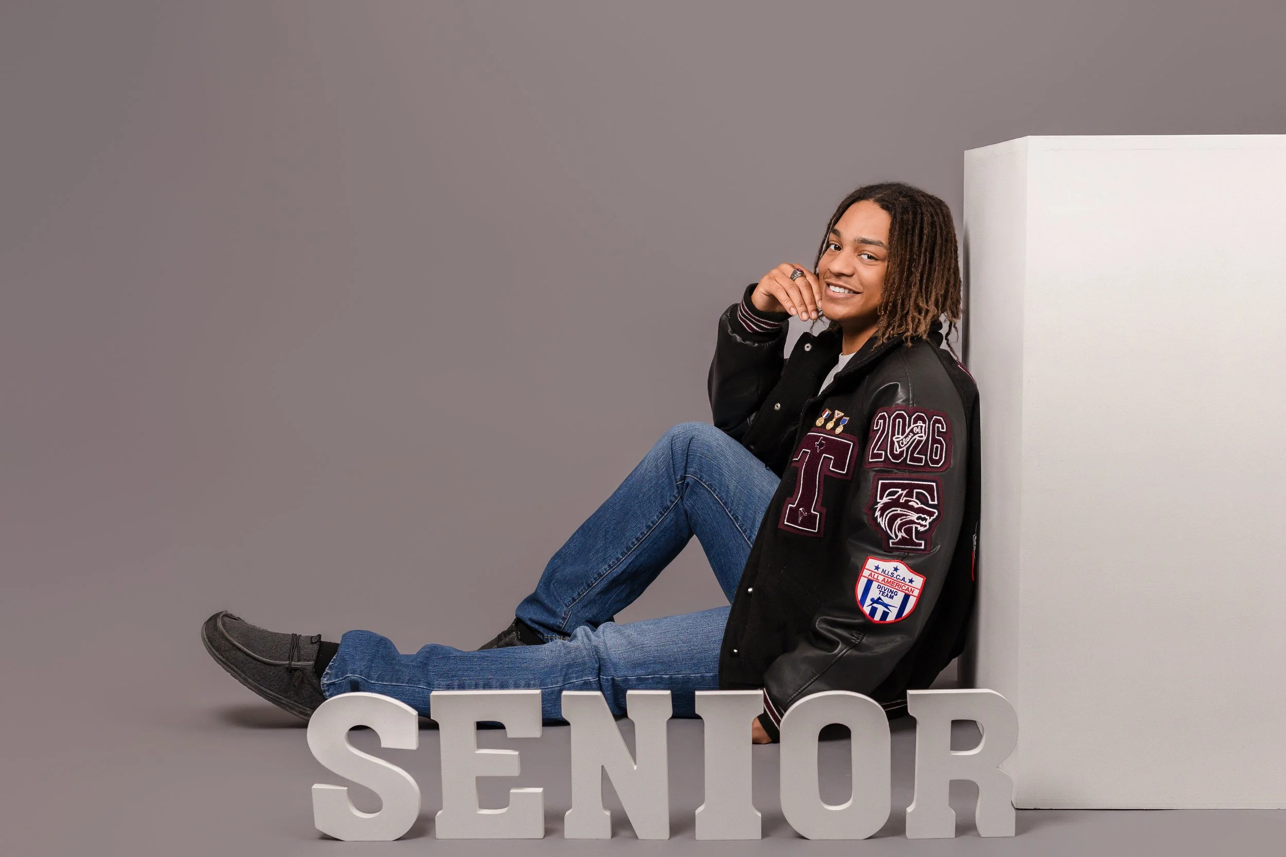 Young person with dreadlocks sitting on the floor against a gray background, smiling with one hand near their face, wearing a black varsity jacket with patches and the year 2026, blue jeans, and black shoes, with large white 3D letters spelling 'SENI