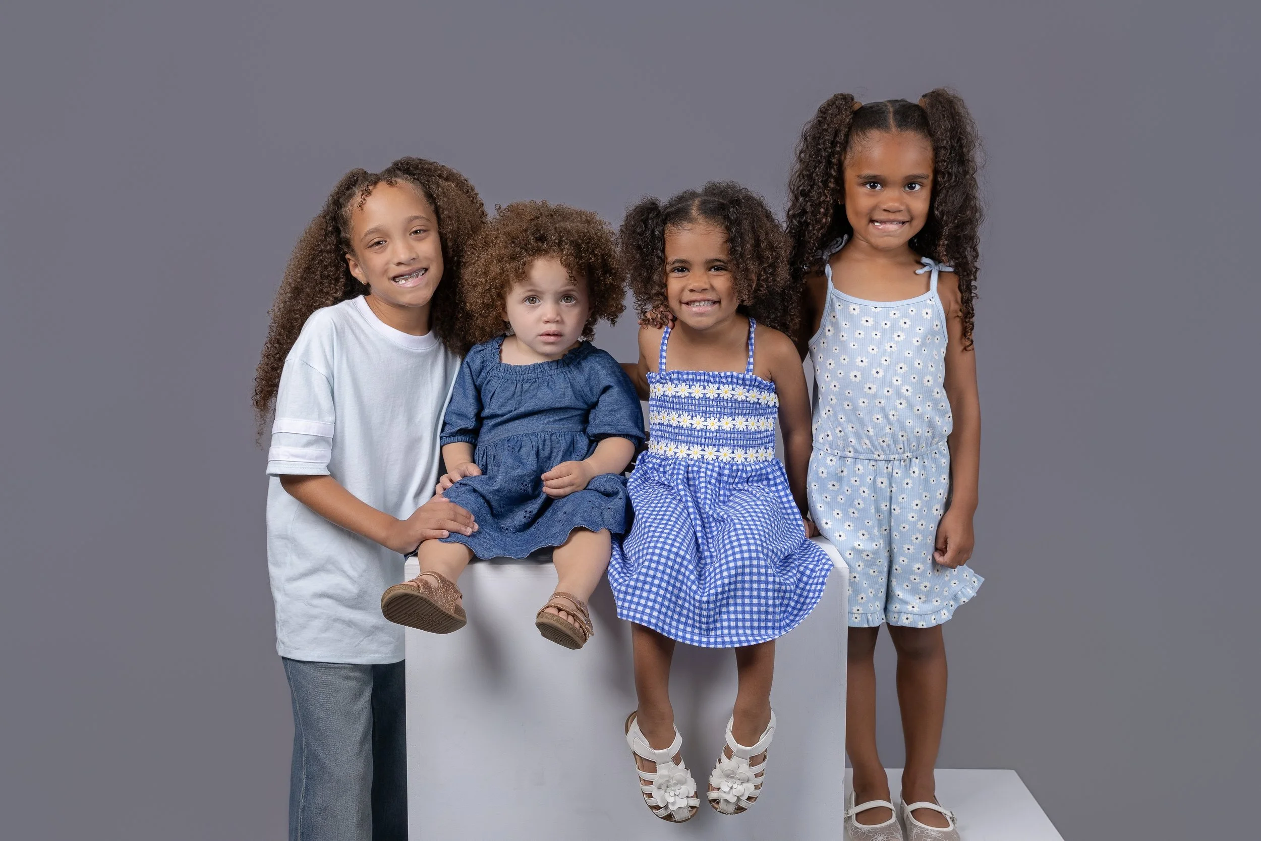 Four sibling girls smiling together during a studio portrait session in Burleson, TX, with the youngest child seated on a stool and the older children gathered around against a gray backdrop.