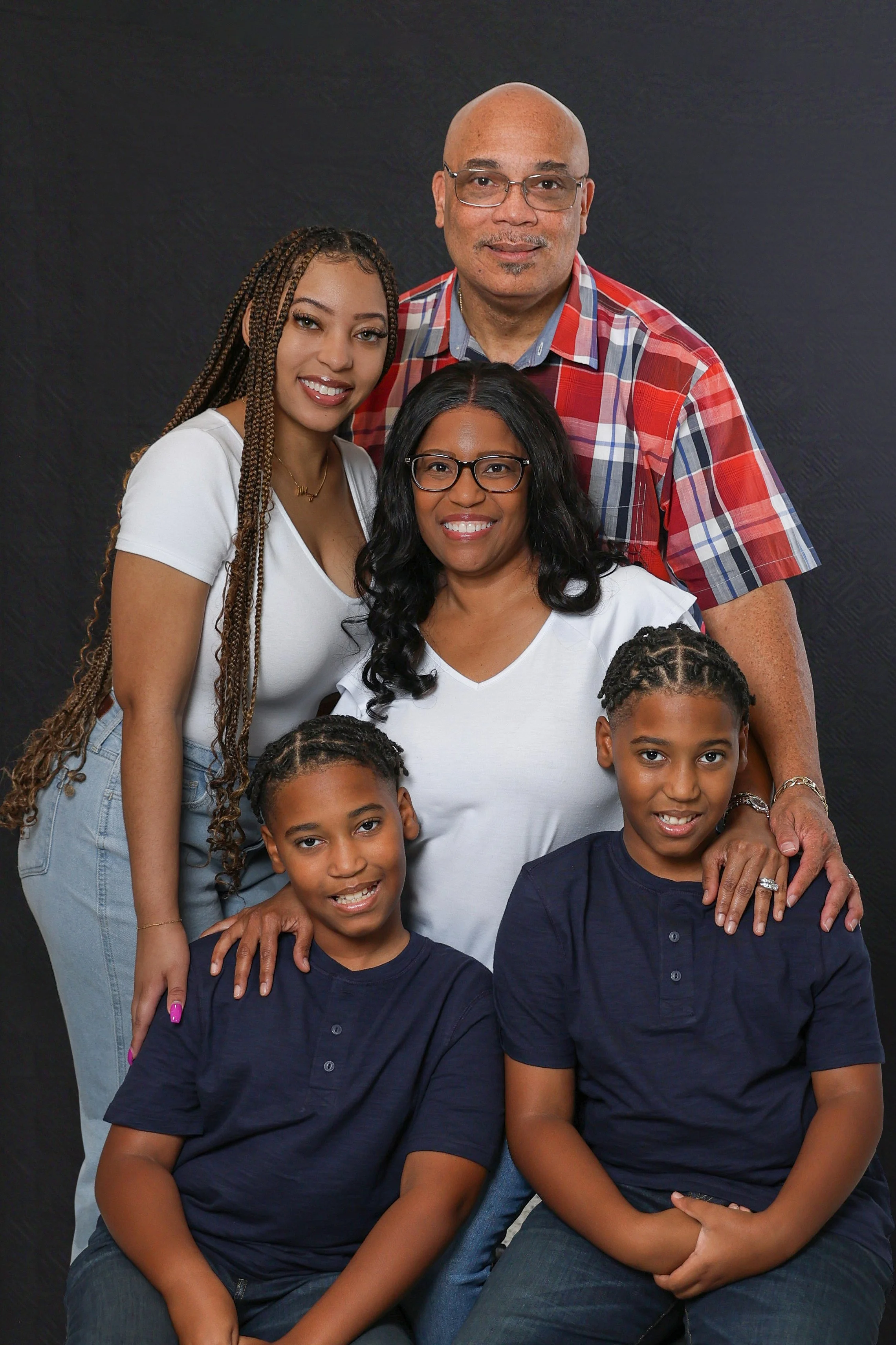 A family portrait of four members against a black background. An older man with glasses and a bald head in a red plaid shirt stands behind a smiling woman with glasses and long black hair. Two young boys with dark hair and blue shirts sit in front, s