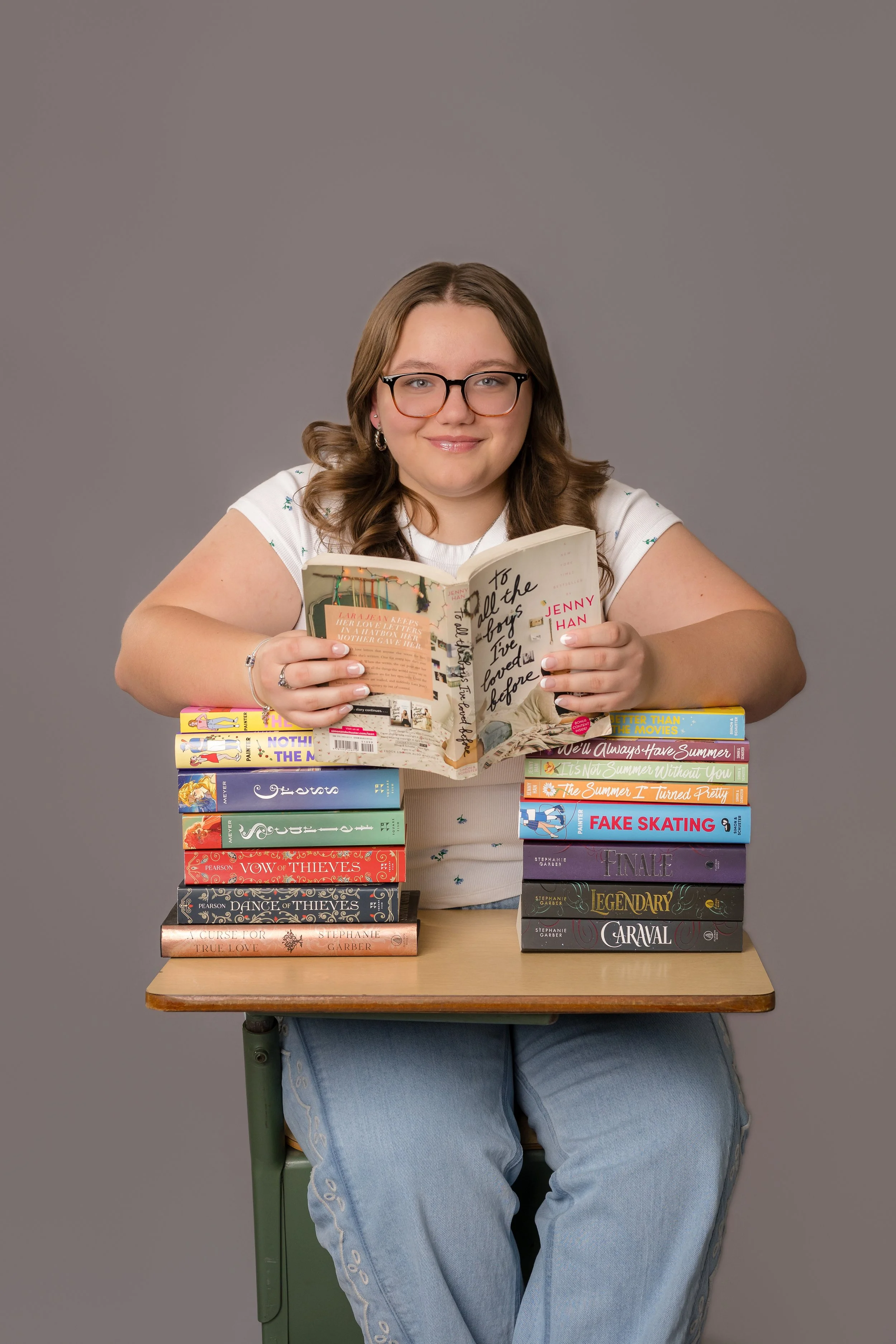 A young woman with glasses and long, wavy brown hair is sitting at a desk, smiling and holding an open book. She is surrounded by a stack of colorful books on the desk. The background is plain gray.