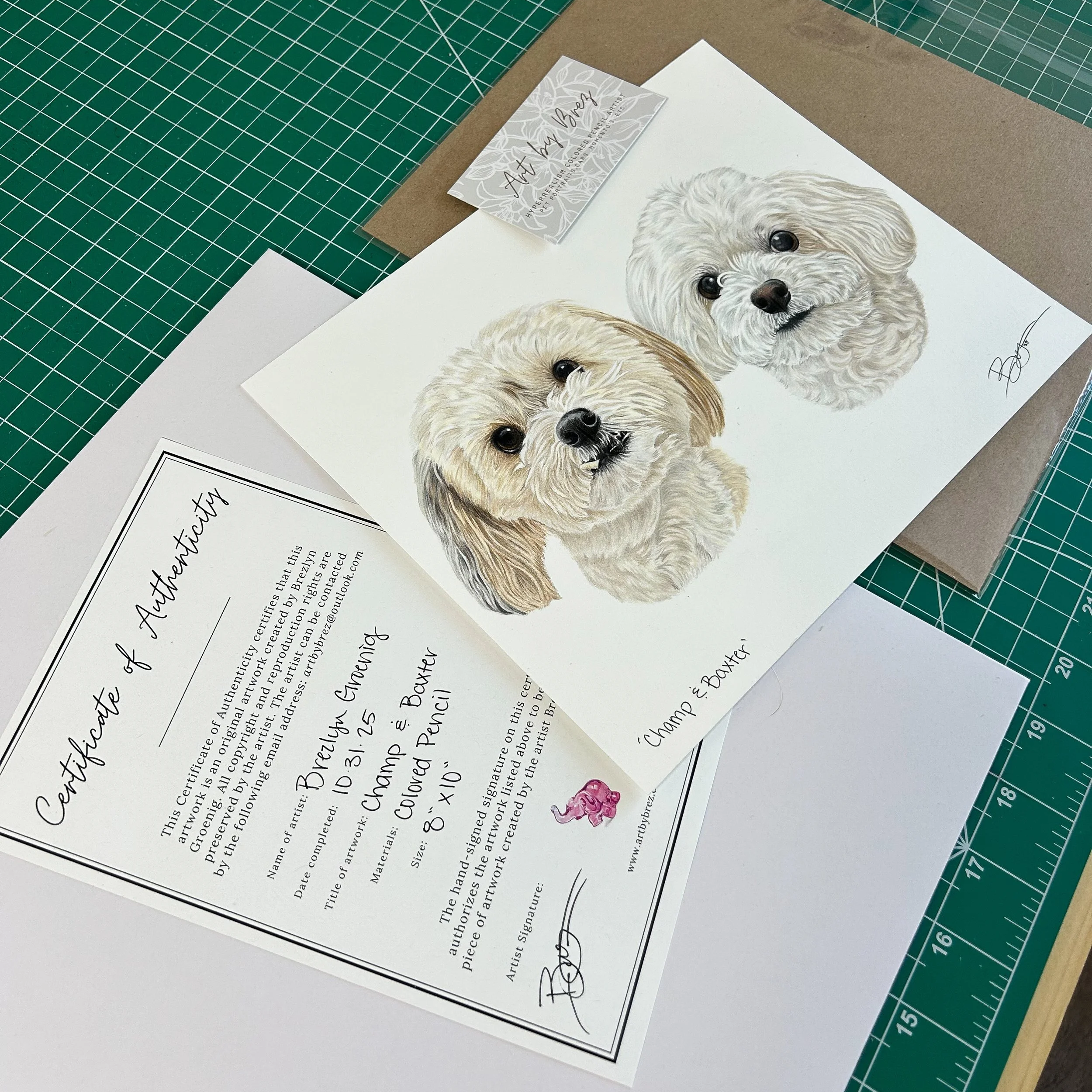 Art print of two illustrated dogs with signatures, accompanied by a certification of authenticity and a business card for ArbyBree. The art is on a white sheet on a green cutting mat with a ruler, and the certification is on white paper.