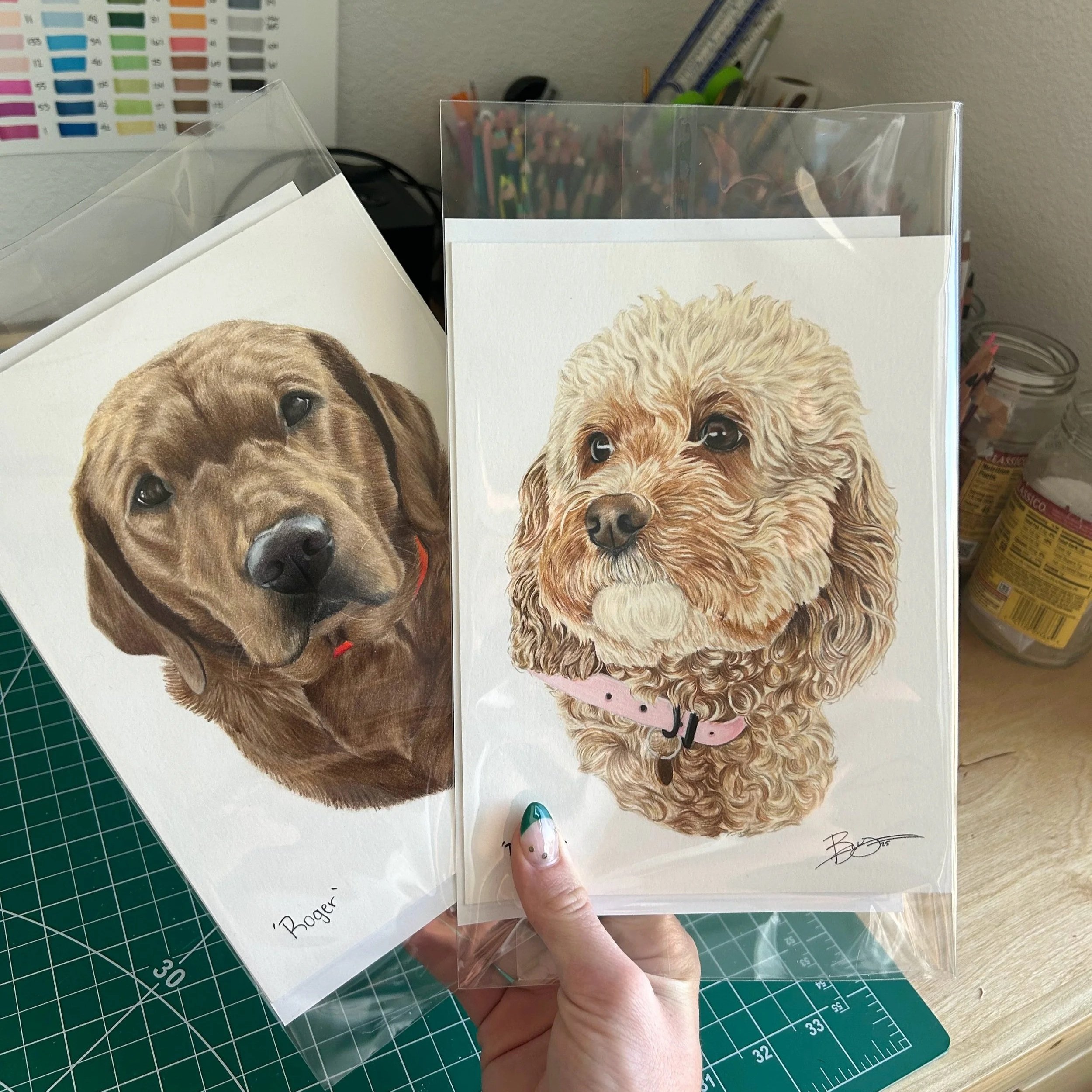 Two illustrated dog portraits in clear plastic sleeves. One is a brown Labrador retriever with a red collar, the other is a curly-haired beige dog with a pink collar. The background shows a crafting workspace with cutting mat, paint color chart, and 