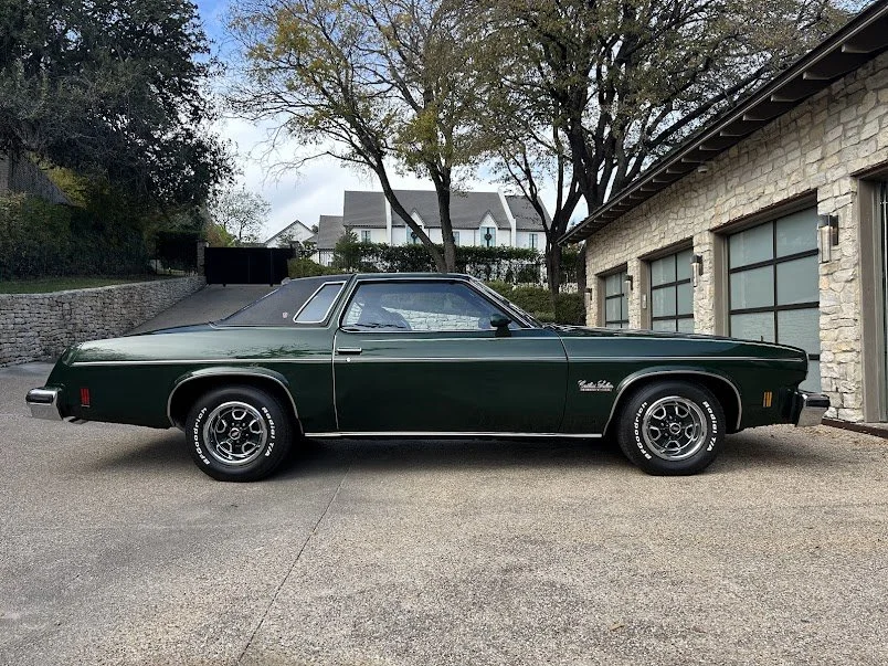 A dark green vintage Oldsmobile Cutlass car parked on a driveway in front of a stone house with garage doors. Trees are visible in the background.