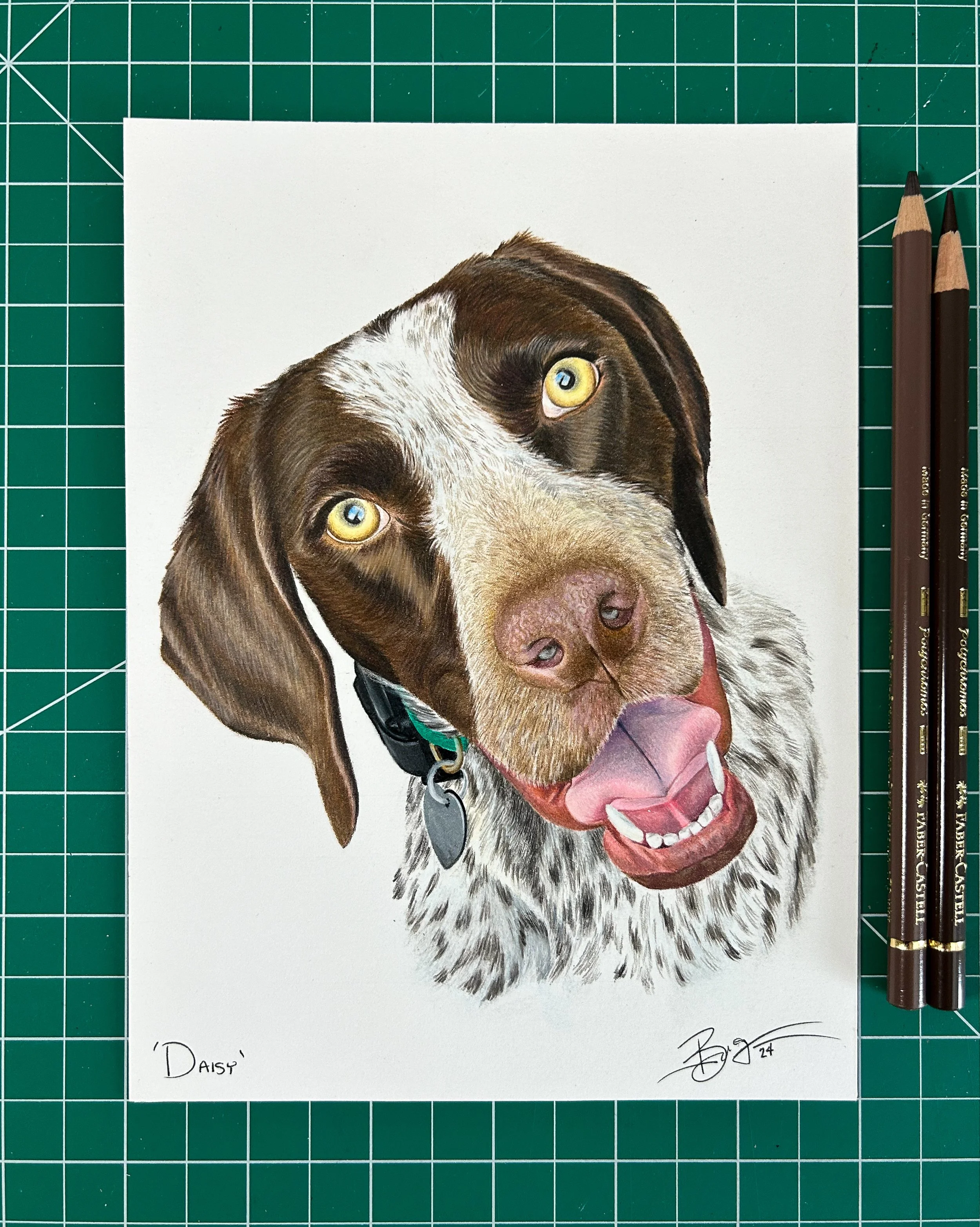 Colored pencil drawing of a happy brown and white dog with yellow eyes, wearing a collar with tags, on white paper placed on a green cutting mat with grid lines.