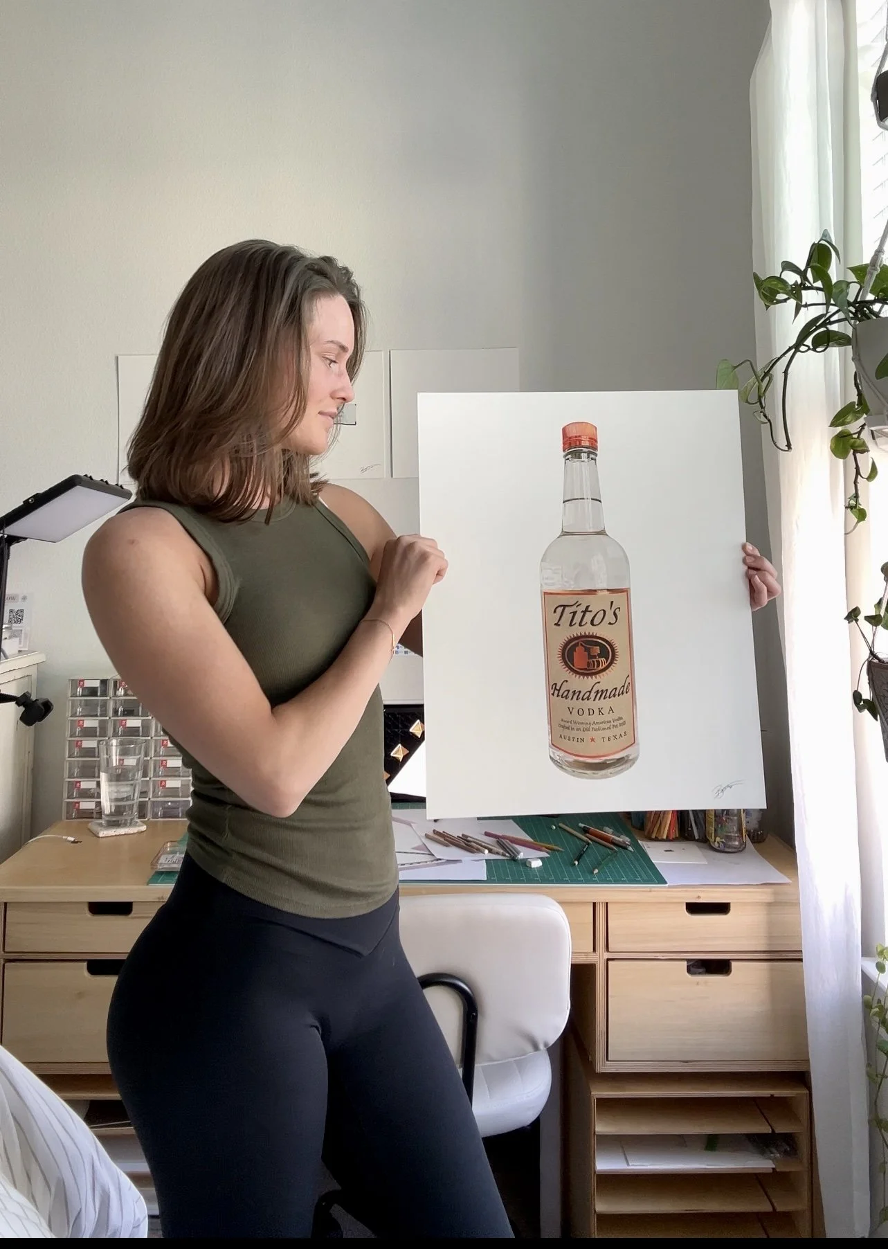 A woman holding a large painting of a bottle of Tito's Handmade Vodka in a bright room with a desk, art supplies, and plants.