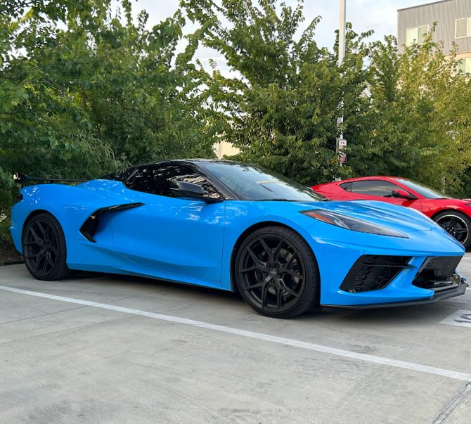 Blue sports car parked in a lot with a red sports car behind it and trees in the background.