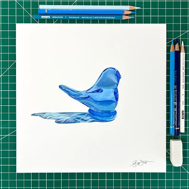 A realistic drawing of a blue glass shoe placed on white paper, with colored pencils and an eraser around it on a green cutting mat.