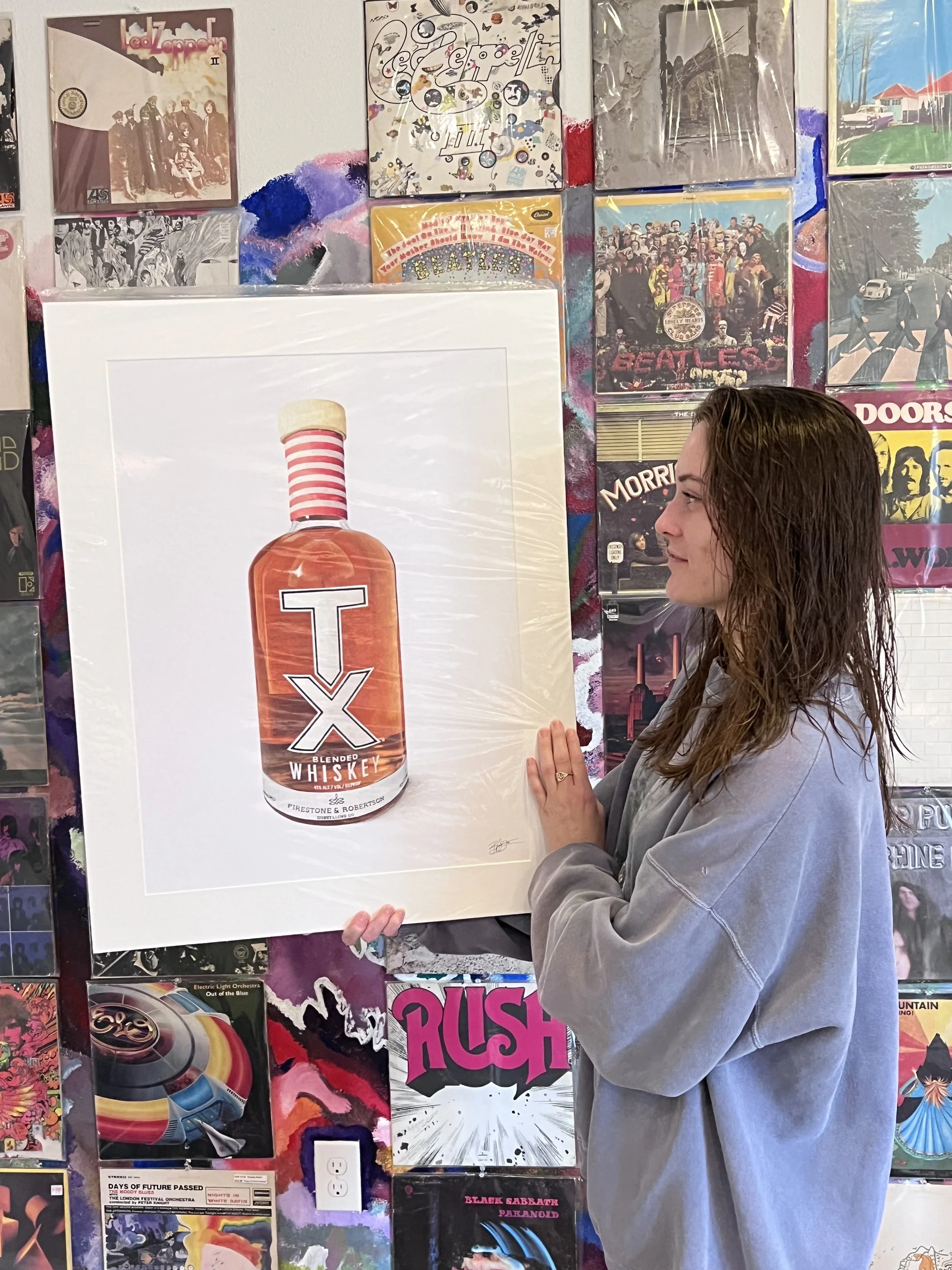 A woman holding a poster of Firestone whiskey bottle in front of a colorful wall decorated with various music album covers.